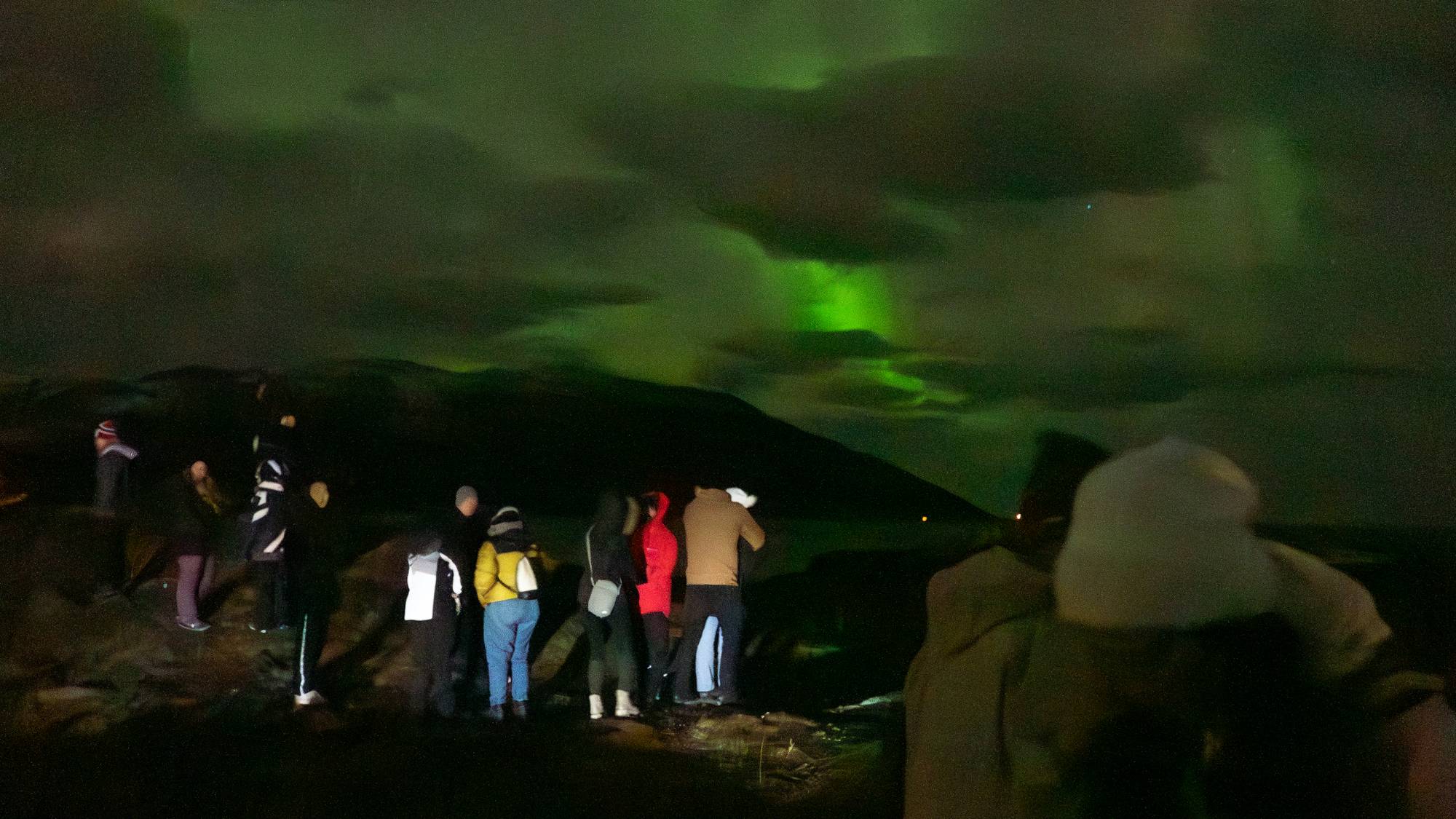 Turister står og ser på nordlys, og tar bilde av det. Himmelen lyser opp av grønt nordlys
