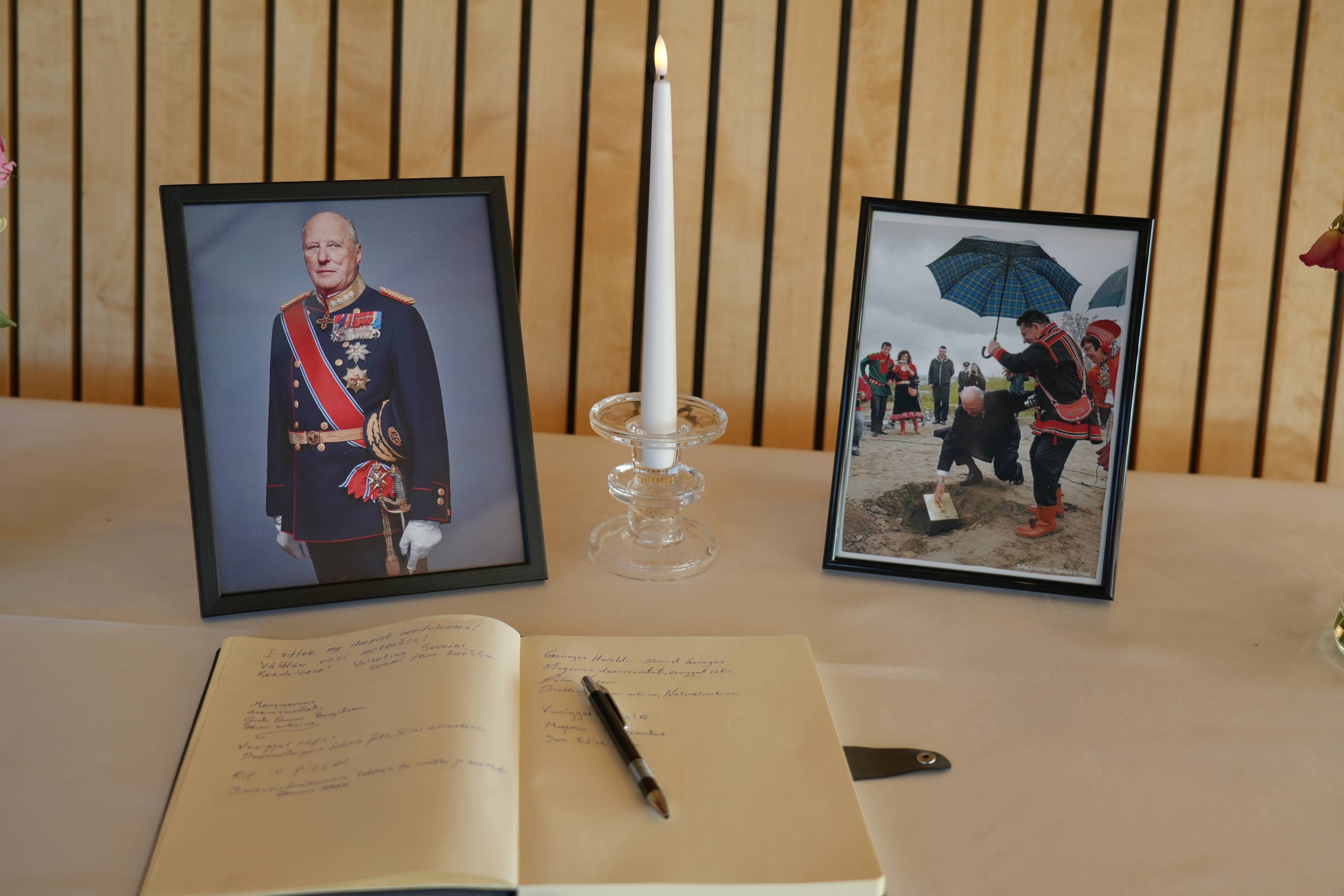 Kondolanseprotokoll, levende lys og to innrammede fotografier av Kong Harald på et bord. (Bildebeskrivelsen er laget av en KI-tjeneste)