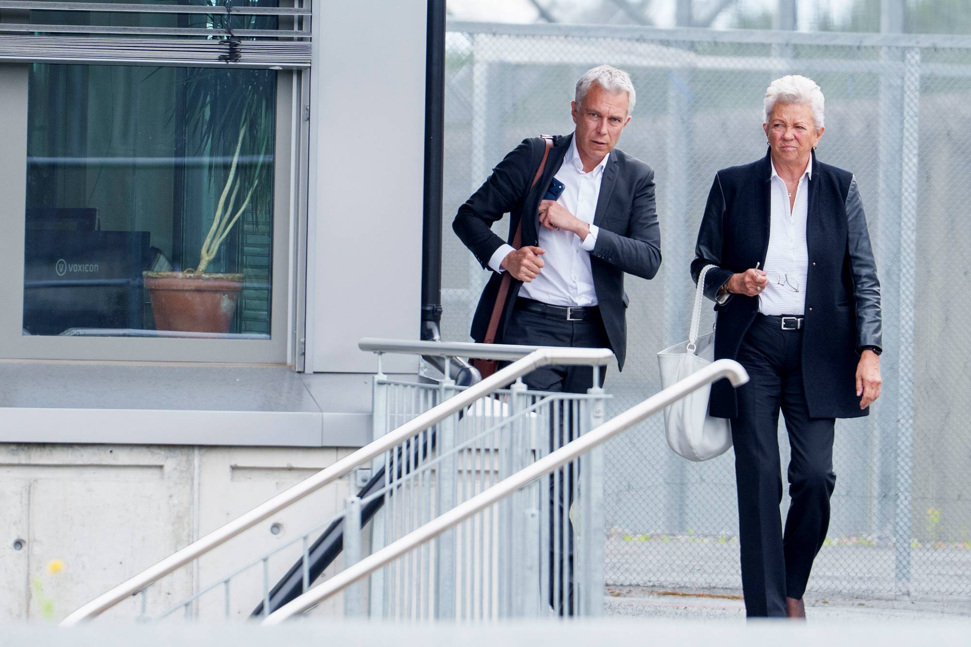 Mann og kvinne i formelle klær går ut av en bygning ved et inngjerdet område. (Bildebeskrivelsen er laget av en KI-tjeneste)