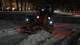 Photo : Bard Nafstad / NRK Une machine de déneigement travaille dans l’obscurité de la nuit pour déneiger la route. La machine est équipée d'une grande charrue à l'avant qui repousse actuellement la neige. Les environs montrent des rues et des bâtiments enneigés, avec des lumières tamisées provenant des zones environnantes. Le temps est nuageux et la neige recouvre le sol d'une épaisse couche. (La description de l'image est créée par un service AI)