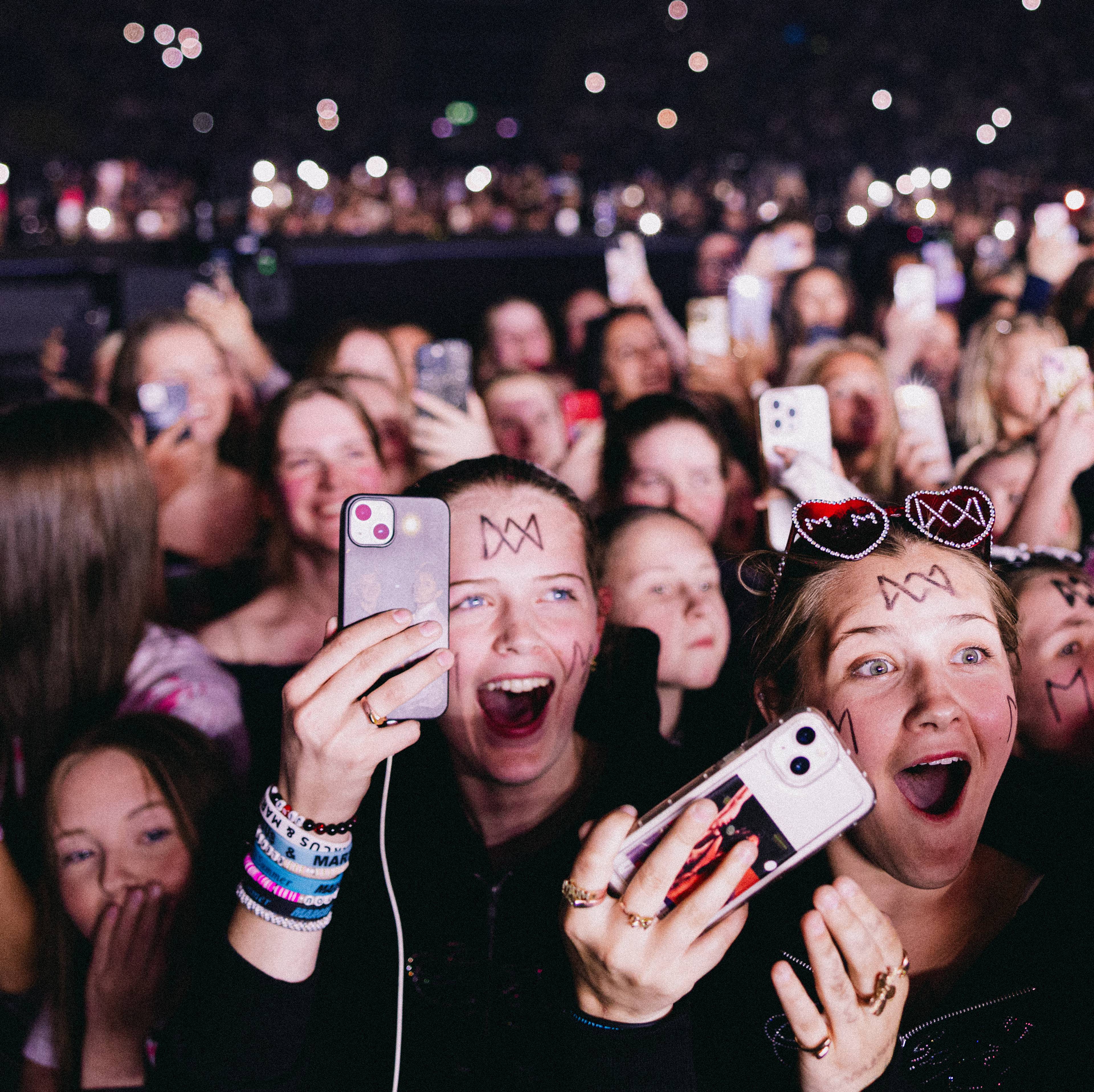 Publikum holder opp mobiltelefoner under en konsert. Flere unge kvinner med ansiktsmaling og hodebånd ser mot scenen. Bak dem er en stor mengde mennesker i et mørkt rom opplyst av mobilskjermer. (Bildebeskrivelsen er laget av en KI-tjeneste)