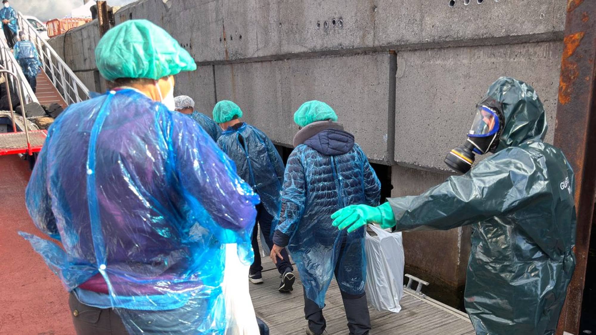 Flere personer i verneutstyr går langs en kai. En person i mørkegrønn fullskjermdrakt med gassmaske leder an. 