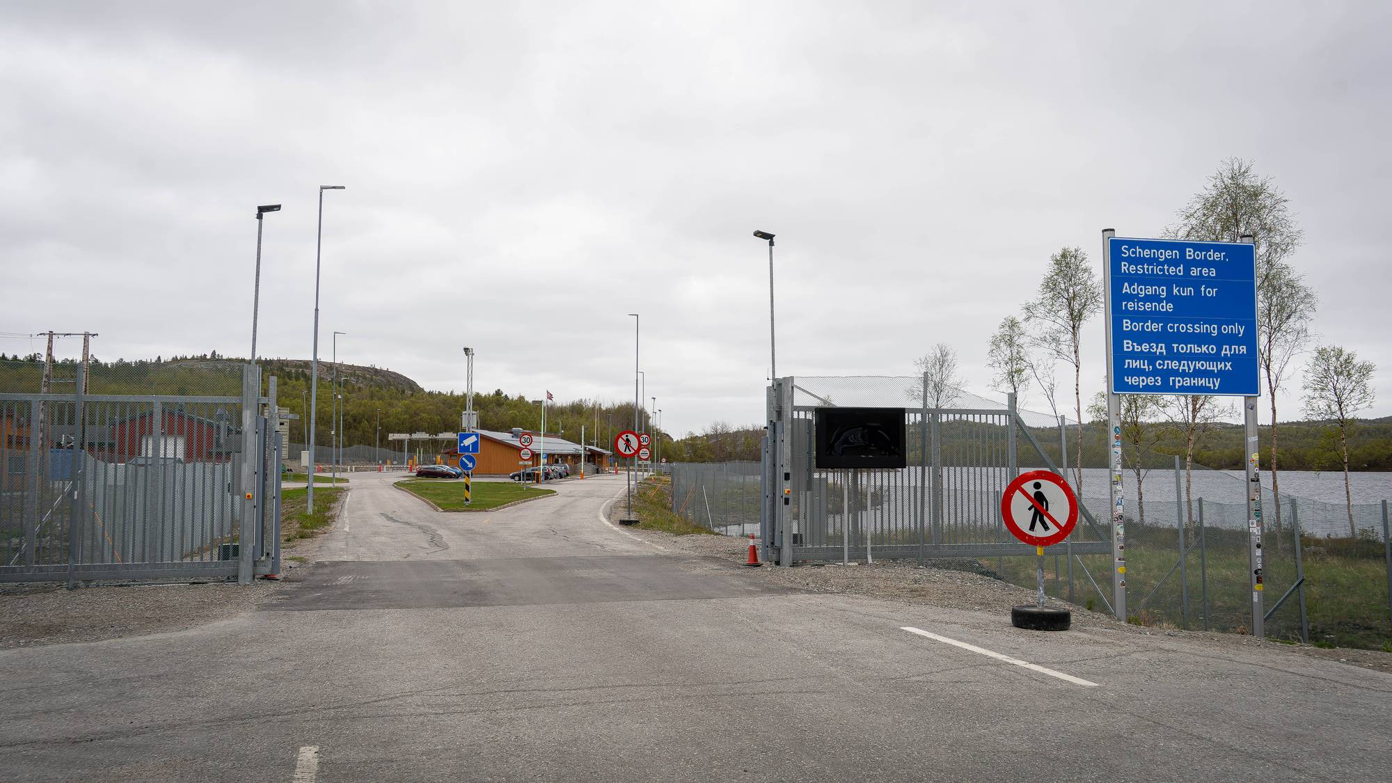 Bildet viser en grensekontrollstasjon med skilt på flere språk. Et blått skilt informerer om Schengen-grensen og at det er et restriktivt område. På en annen stolpe er et rundt skilt med et bilde av en person som krysser en strek. Veien fører mot en bygning med metallporter. (Bildebeskrivelsen er laget av en KI-tjeneste)