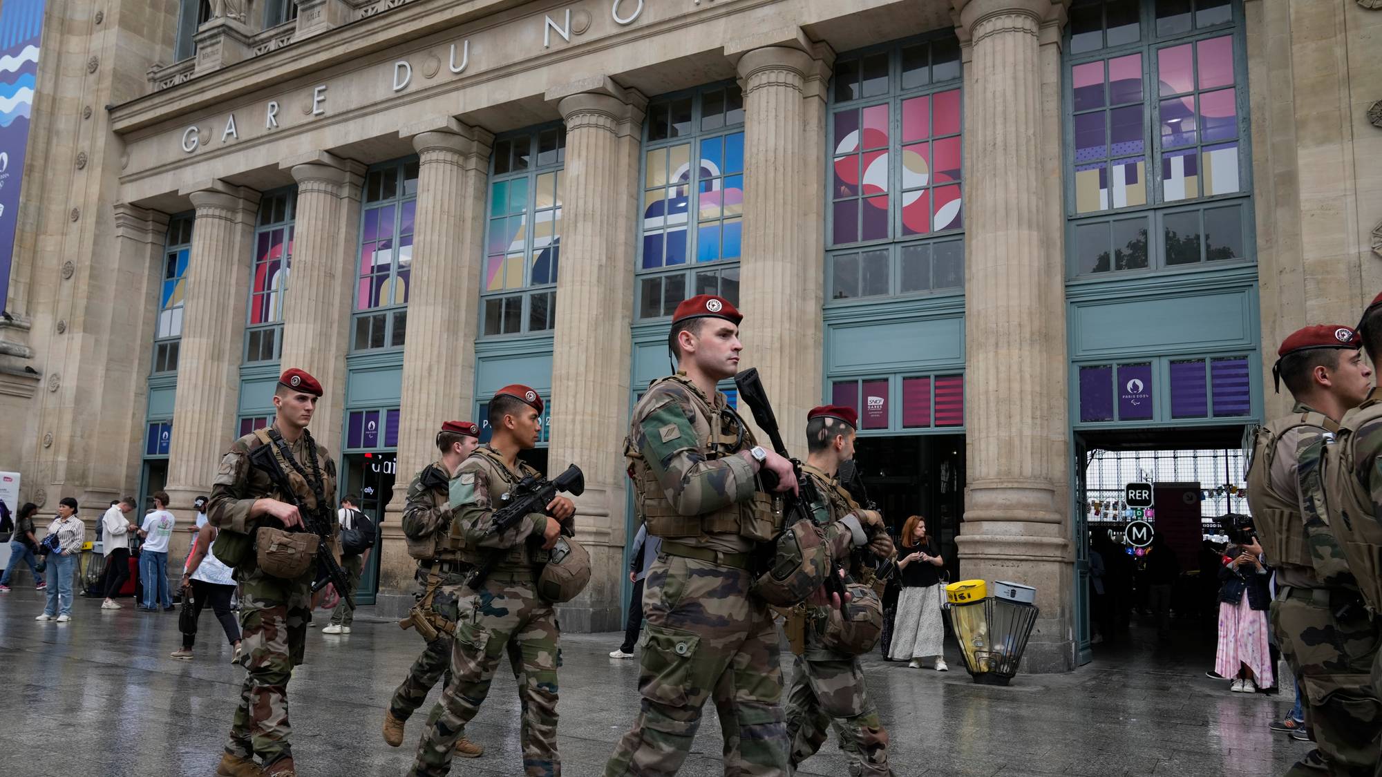 Soldater i kamuflasjeuniform og rød beret patruljerer foran Gare du Nord i Paris. De bærer rifler og annet militært utstyr. Bildet viser en rekke mennesker og bygninger. (Bildebeskrivelsen er laget av en KI-tjeneste)