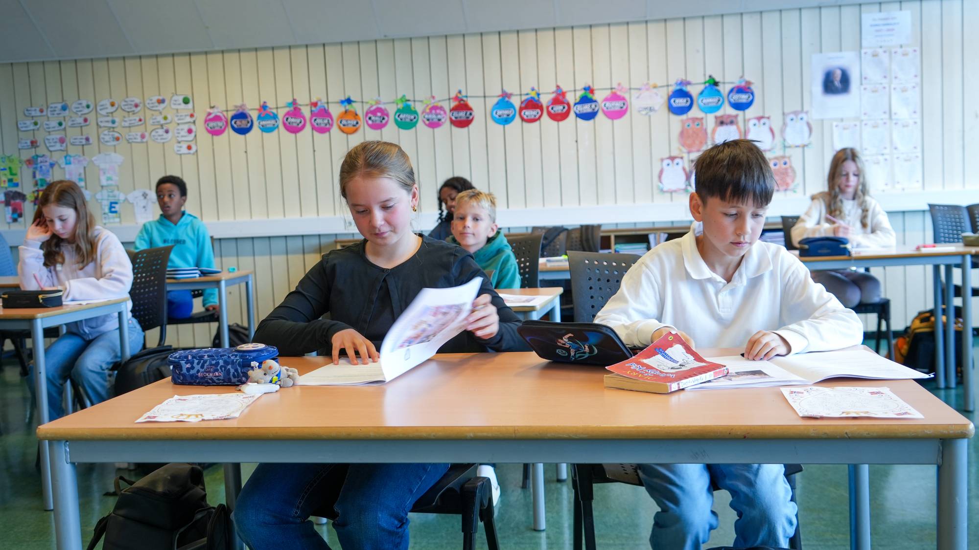 En gutt med mørkt hår og hvit genser og ei jente med lyst hår og svart genser sitter ved siden av hverandre på ved en pult i et klasserom med flere elever bak seg.