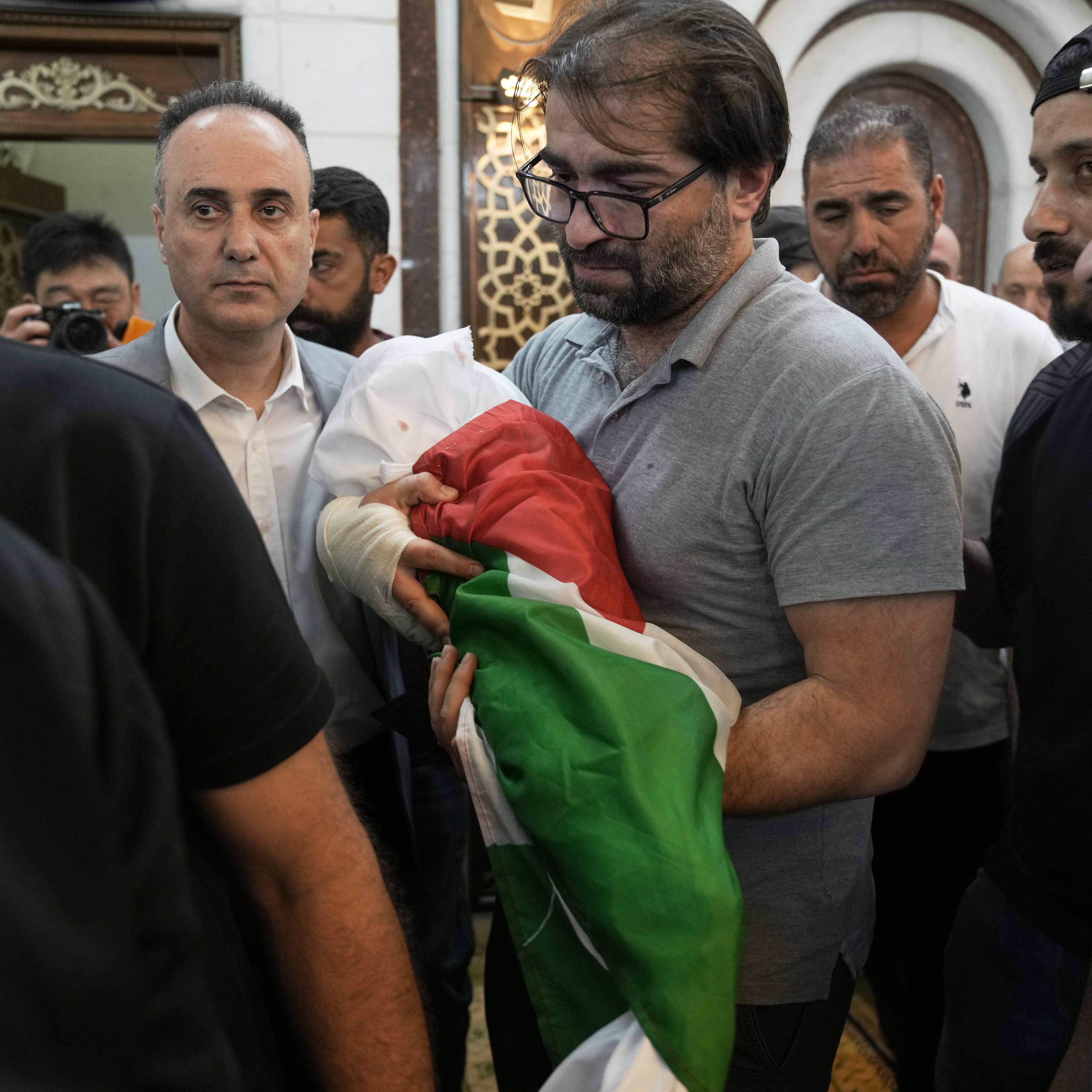 En mann med briller holder et spedbarn pakket inn i det palestinske flagget.Flere menn står rundt og ser på.Bakgrunnen viser et interiør med lyse vegger og et speil.Bildet er tatt innendørs.