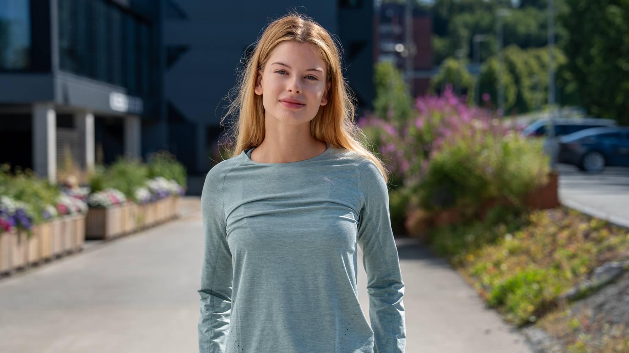 En ung kvinne med langt blondt hår står utendørs i en lys grønn langermet trøye. (Bildebeskrivelsen er laget av en KI-tjeneste)