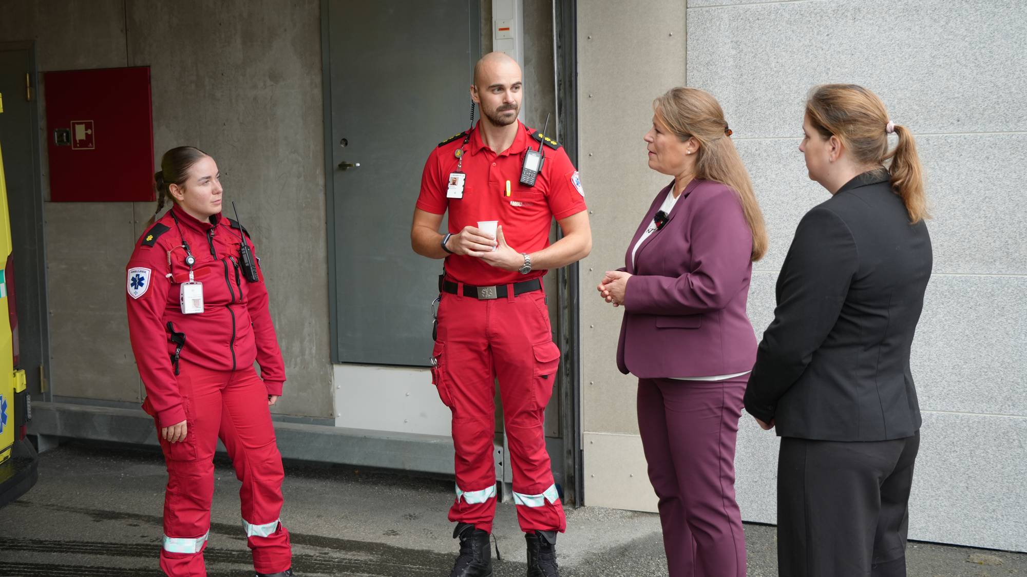 To ambulansearbeidere i uniform snakker med to kvinner i kontorklær foran en vegg. (Bildebeskrivelsen er laget av en KI-tjeneste)