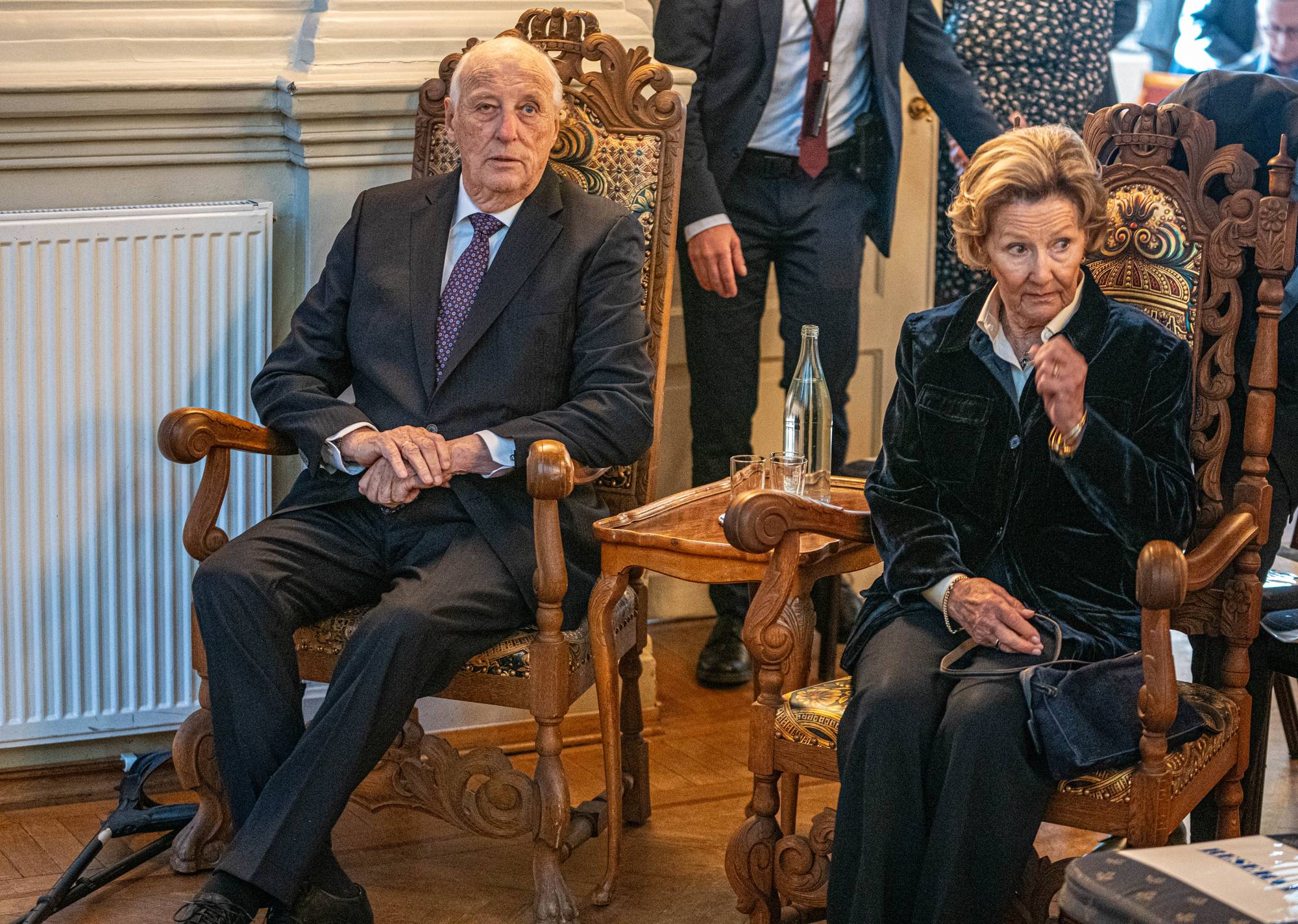 Kong Harald V og dronning Sonja sitter i utsmykkede stoler. Kongen er iført mørk dress og lilla slips, mens dronningen har en mørk jakke. Bak dem står flere personer, og et lite bord med en vannflaske er plassert mellom dem. (Bildebeskrivelsen er laget av en KI-tjeneste)