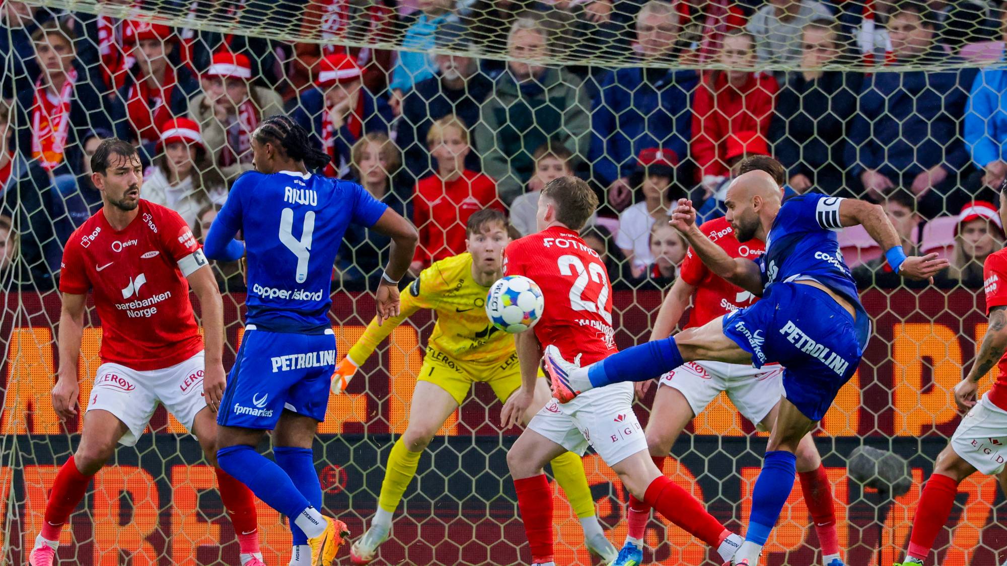 Fotballspillere i røde og blå drakter kjemper om ballen foran målet under en kamp. (Bildebeskrivelsen er laget av en KI-tjeneste)