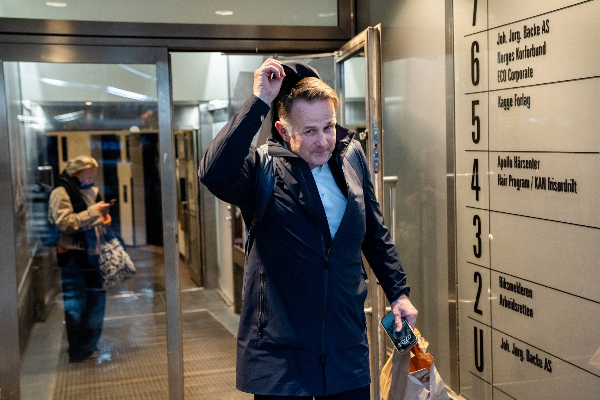 En mann med mørk jakke tar av seg en hette mens han går ut av en dør. Ved siden av er en liste med firmanavn. En annen person ses i bakgrunnen. (Bildebeskrivelsen er laget av en KI-tjeneste)