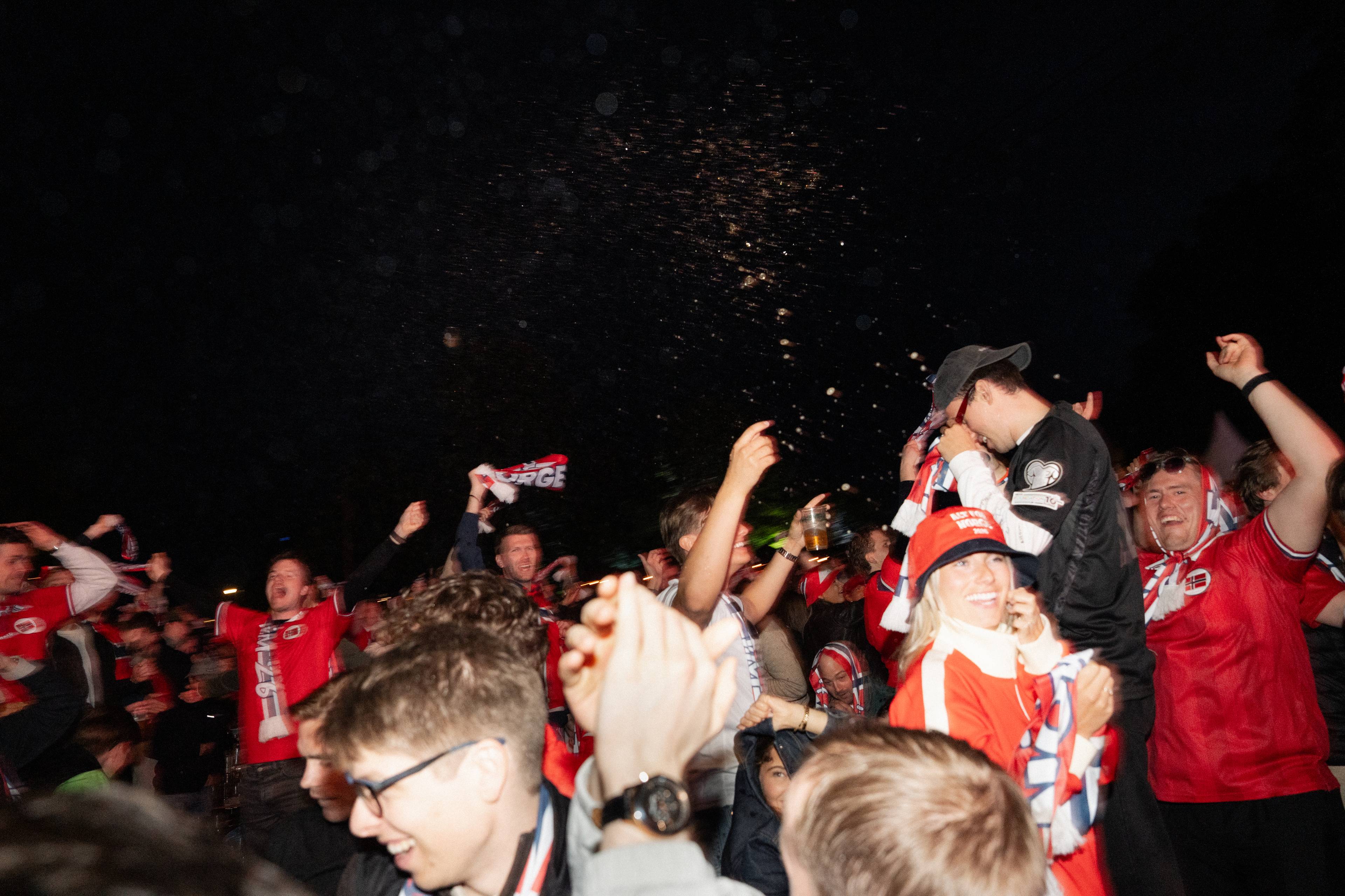 En gruppe mennesker feirer utendørs om natten. Mange bærer supporterutstyr i rødt og hvitt, og flere vifter med flagg. Noen holder opp ølglass. I bakgrunnen er det mørkt med noen lyspunkter. (Bildebeskrivelsen er laget av en KI-tjeneste)