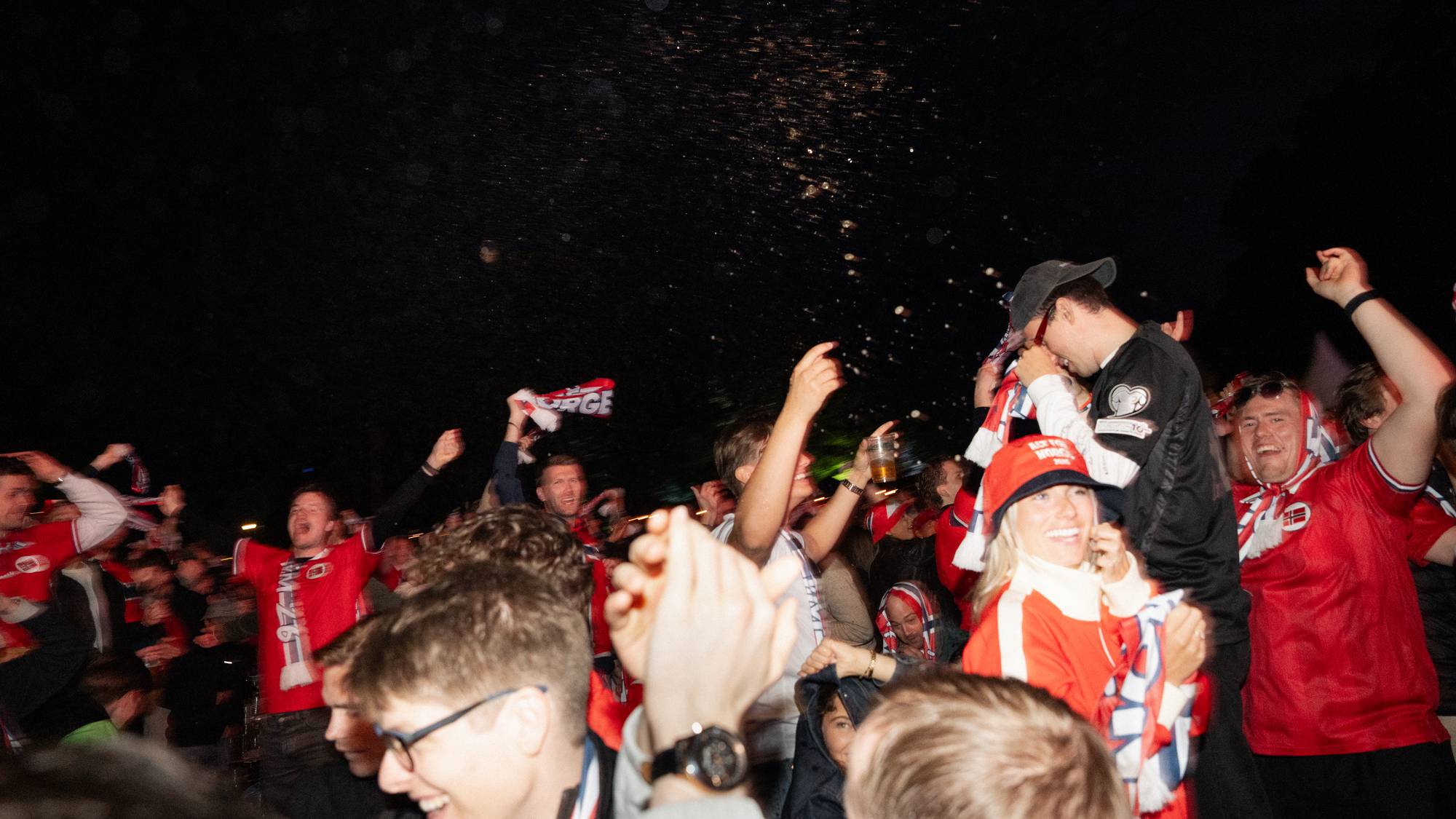 En gruppe mennesker feirer utendørs om natten. Mange bærer supporterutstyr i rødt og hvitt, og flere vifter med flagg. Noen holder opp ølglass. I bakgrunnen er det mørkt med noen lyspunkter. (Bildebeskrivelsen er laget av en KI-tjeneste)