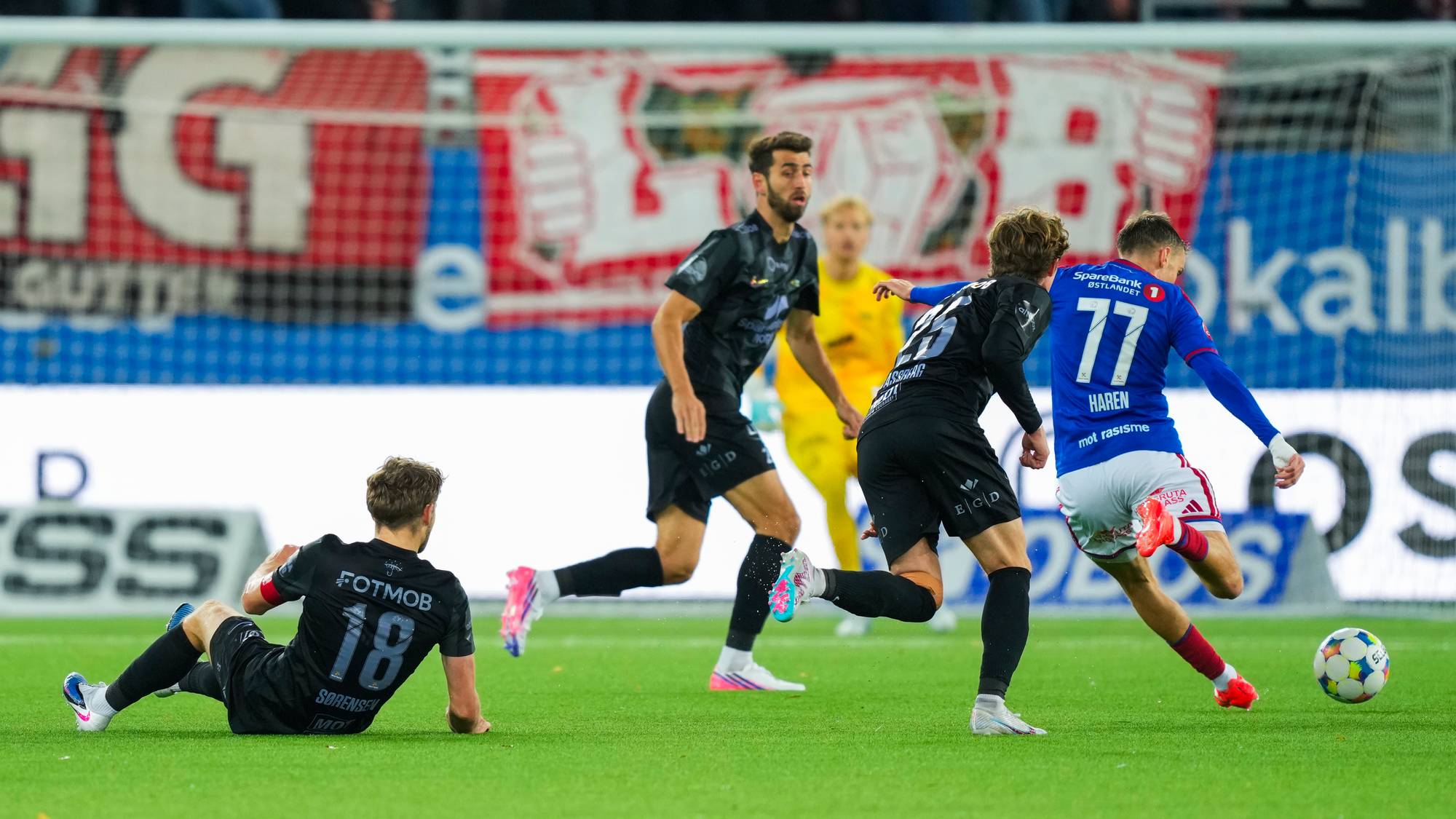 Fotballspiller i blå drakt skyter ballen foran motspillere i svarte drakter på en fotballbane. (Bildebeskrivelsen er laget av en KI-tjeneste)