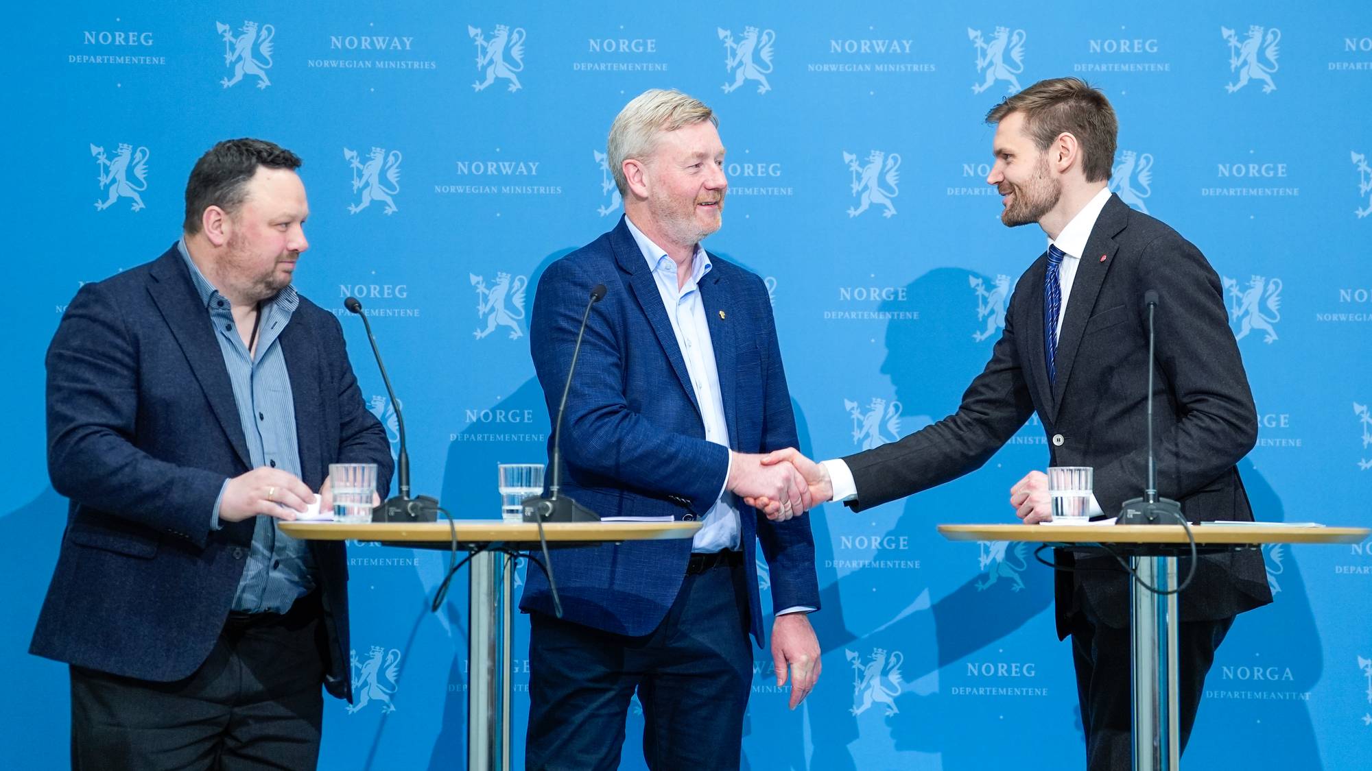 Tre menn står bak et bord foran en blå bakgrunn med Norges riksvåpen og "NORWAY NORWEGIAN MINISTRIES". To menn står ved siden av hverandre og håndhilser, mens en tredje mann står til venstre. (Bildebeskrivelsen er laget av en KI-tjeneste)