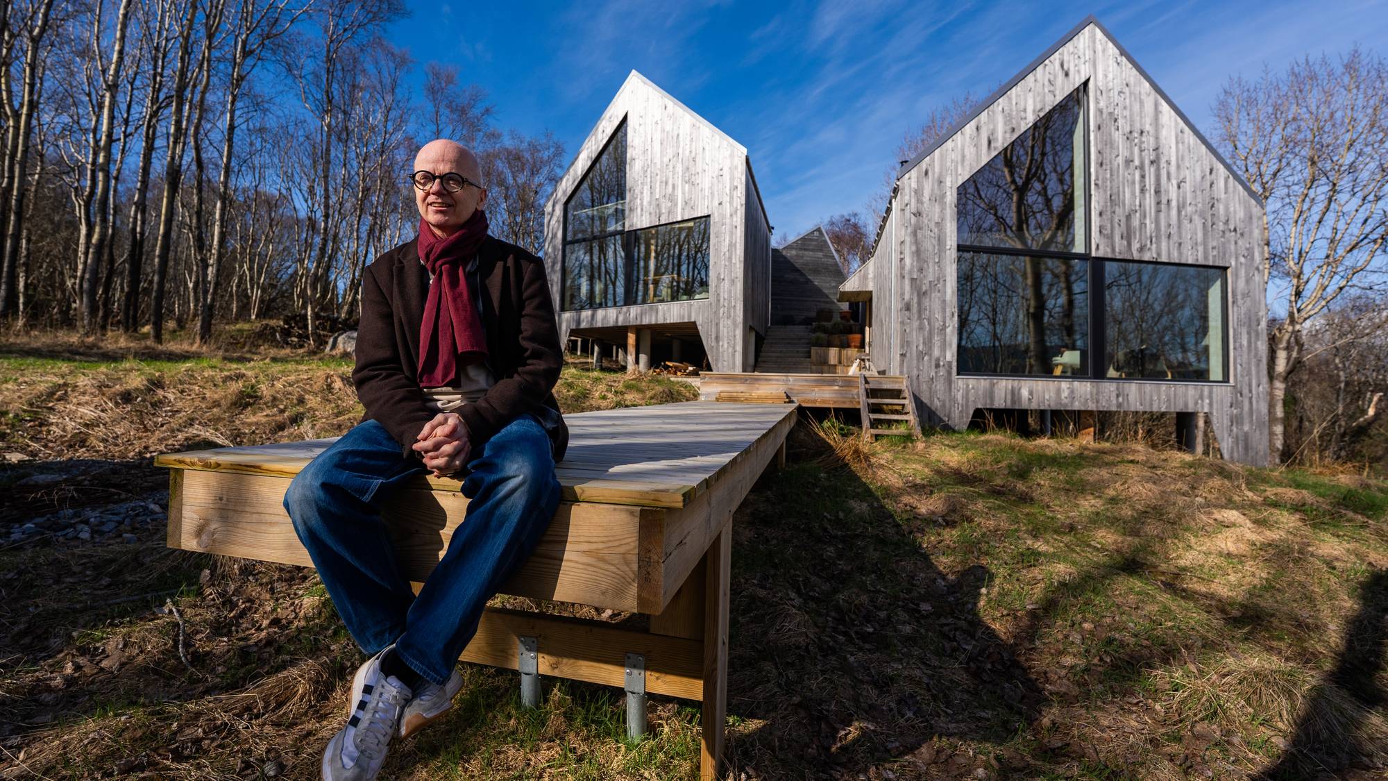 En mann sitter på et utvendig trapperekkverk foran et moderne hus i tre. Huset har et bratt tak og store vinduer. Mannen har på seg en mørk jakke, skjerf og jeans. Solen skinner fra en klar blå himmel. (Bildebeskrivelsen er laget av en KI-tjeneste)
