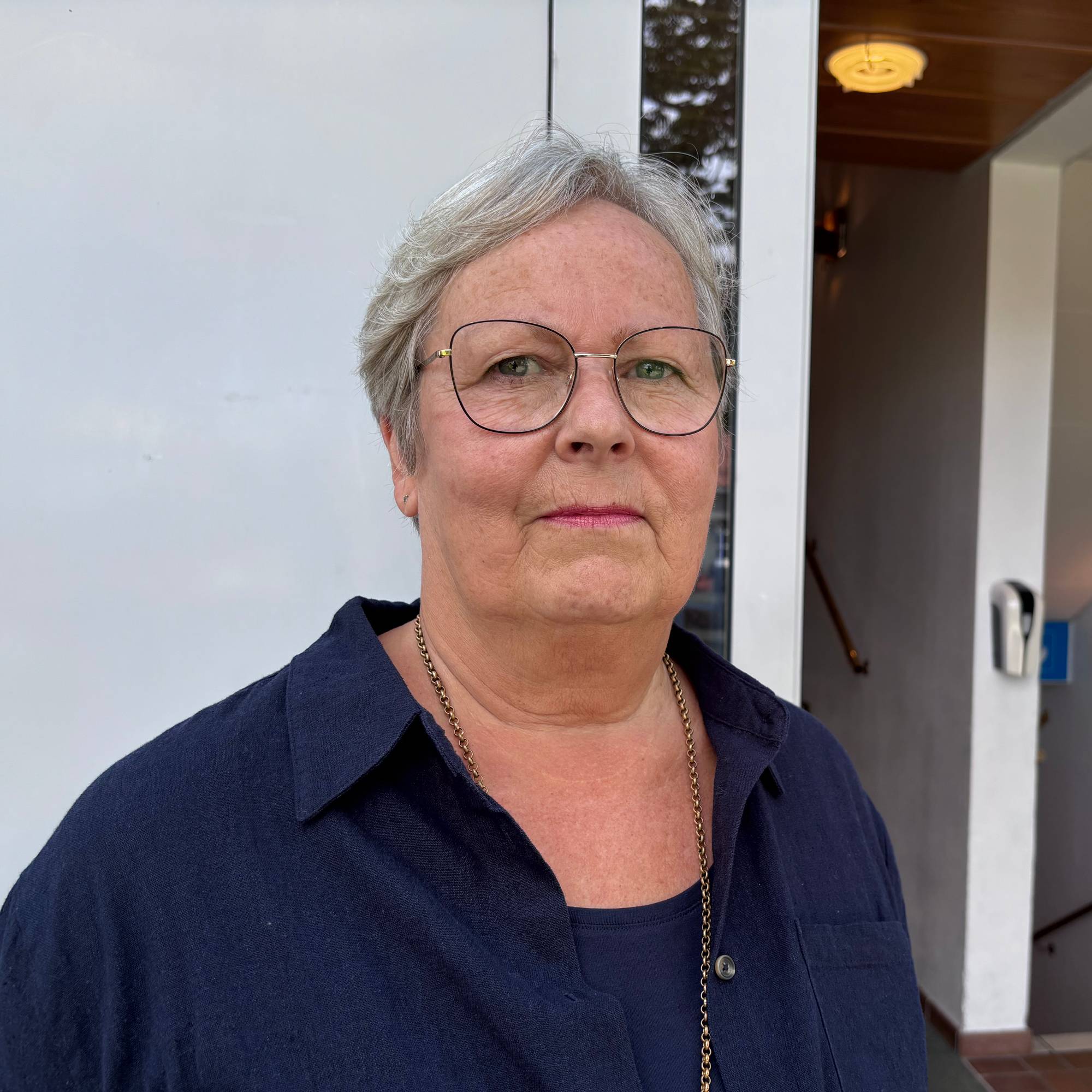En eldre kvinne med briller og grått hår ser inn i kamera. Hun har på seg en mørkeblå skjorte.