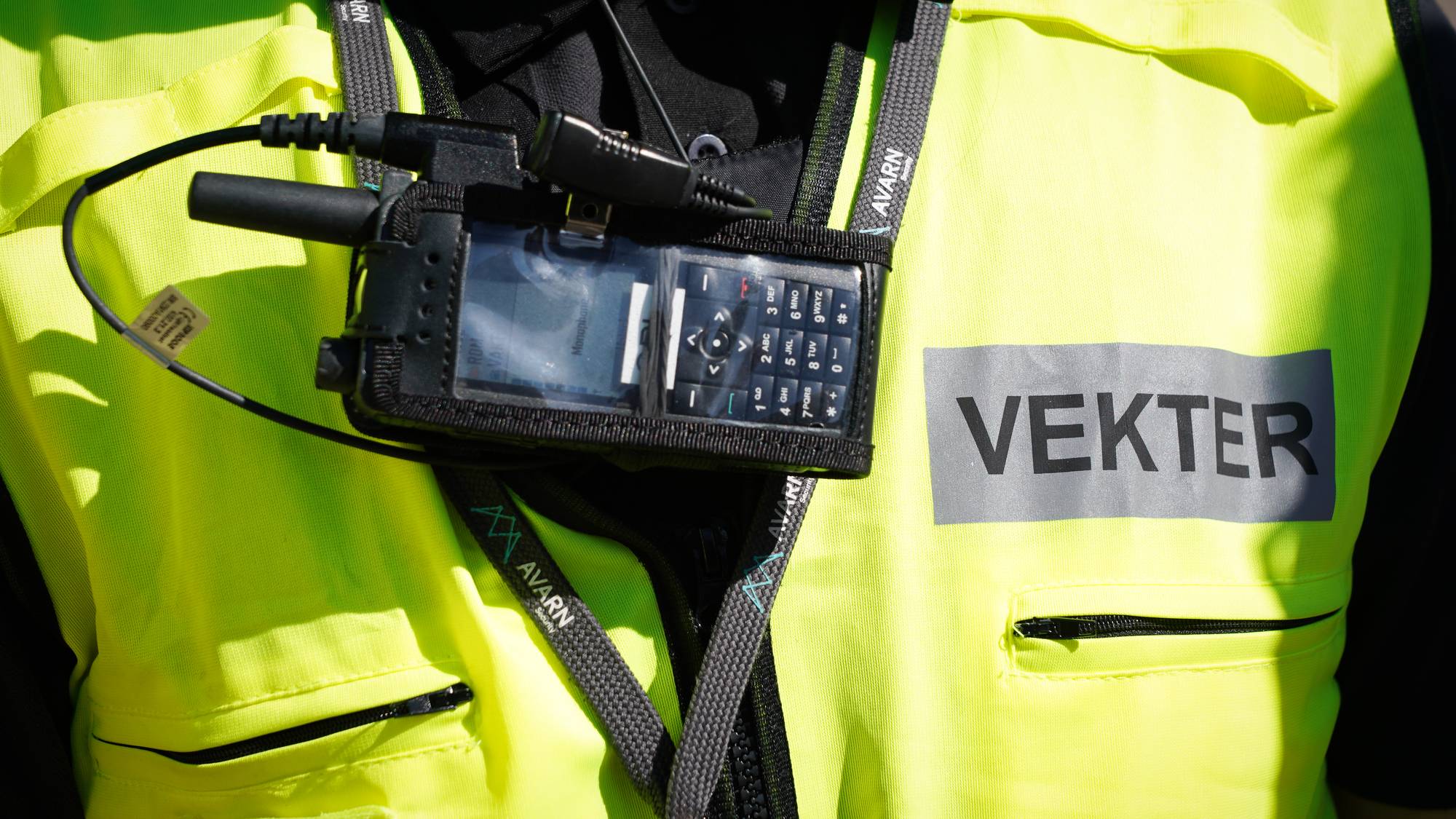 En person med refleksvest har en radiosamband. Ordet "VEKTER" er trykket på vesten. (Bildebeskrivelsen er laget av en KI-tjeneste)