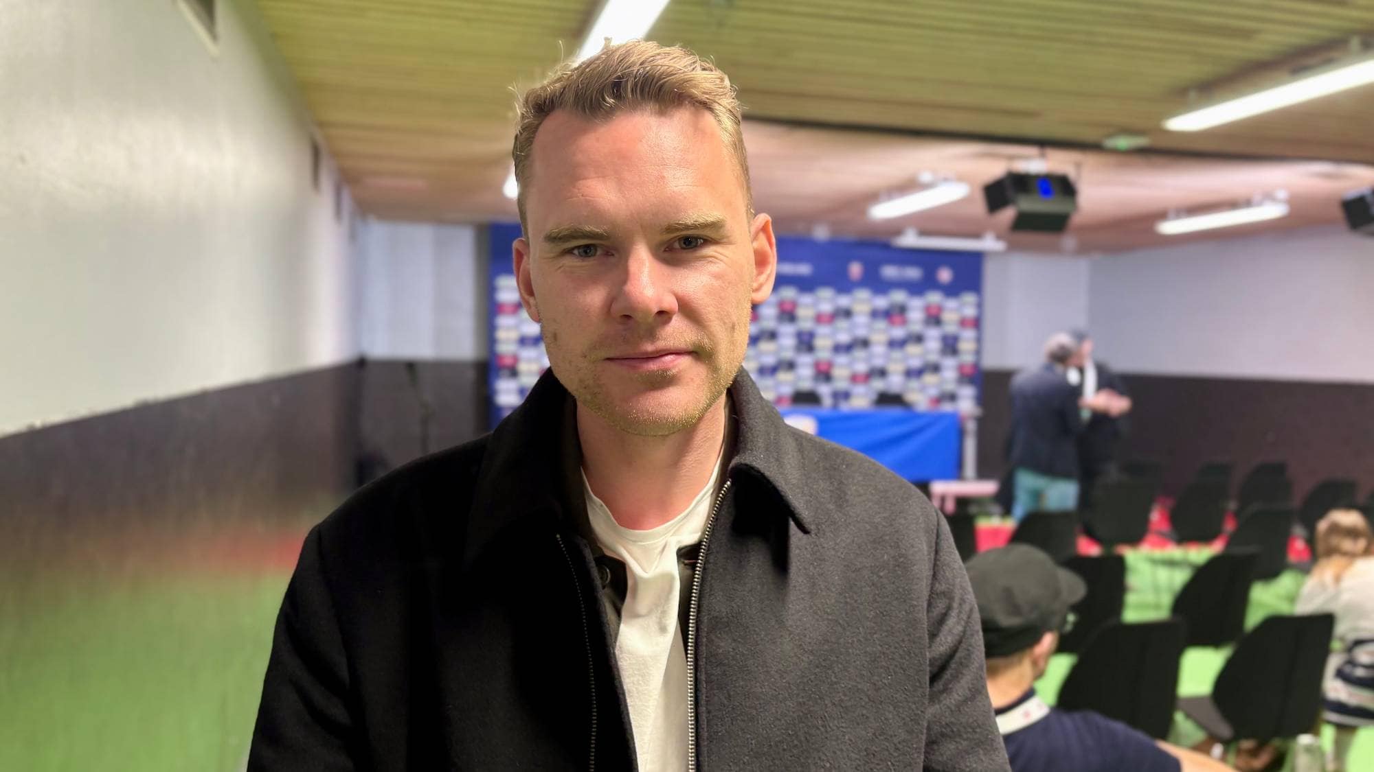 En mann med blondt hår i en mørk jakke står foran et publikum. Bak ham er det et bord dekket med en blå duk og en logo-vegg. Det er flere tomme stoler i rommet. Bakgrunnen er en konferansesal. (Bildebeskrivelsen er laget av en KI-tjeneste)