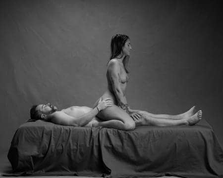 En naken mann med mørkt hår og skjegg ligger på en mørk sofa. En kvinne med tatoveringer på armen og langt mørkt hår sitter skrevende på fanget hans. Hun sitter med ryggen til ham og holder armene på beina hans. Han har armene på hofta hennes.