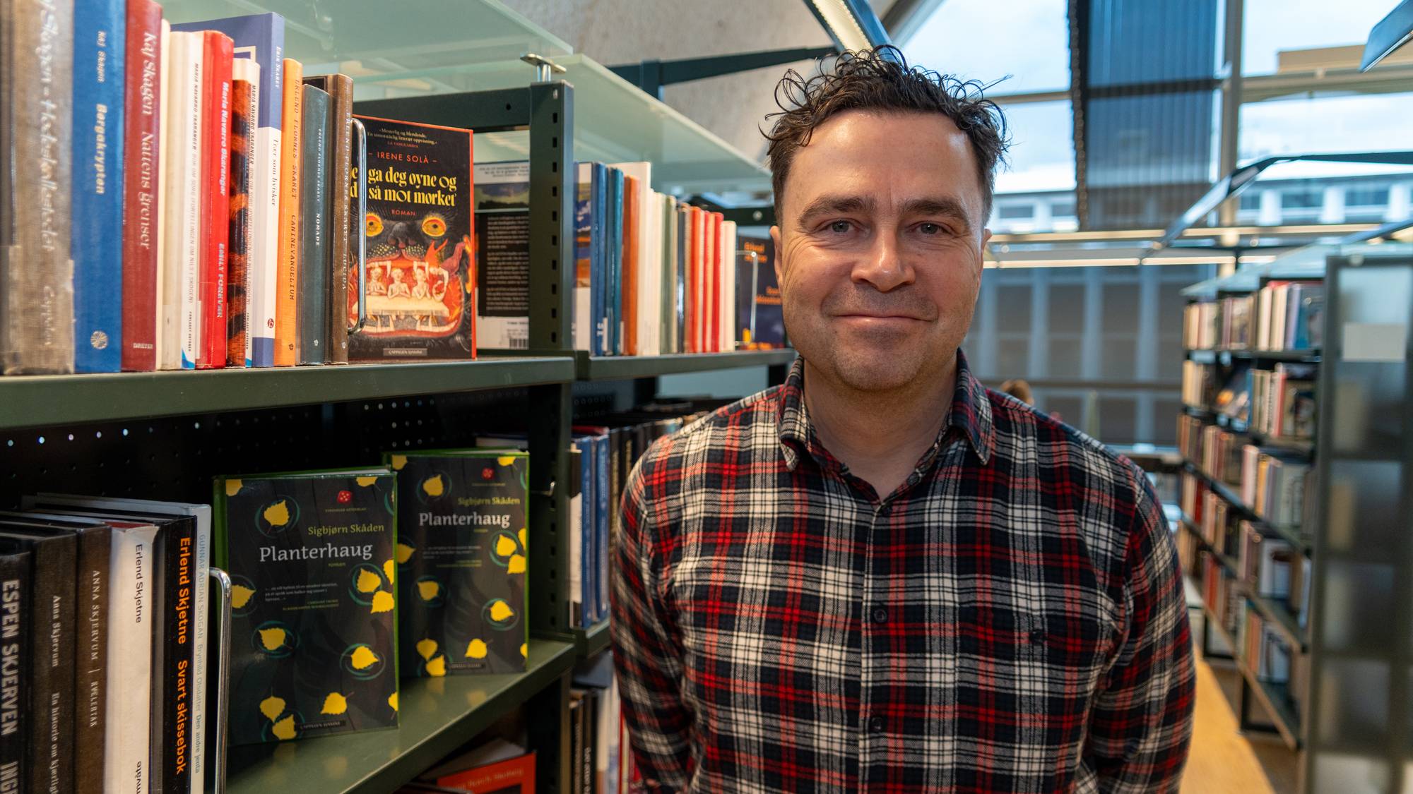 En mann i rutete skjorte står foran en bokhylle i et bibliotek. (Bildebeskrivelsen er laget av en KI-tjeneste)