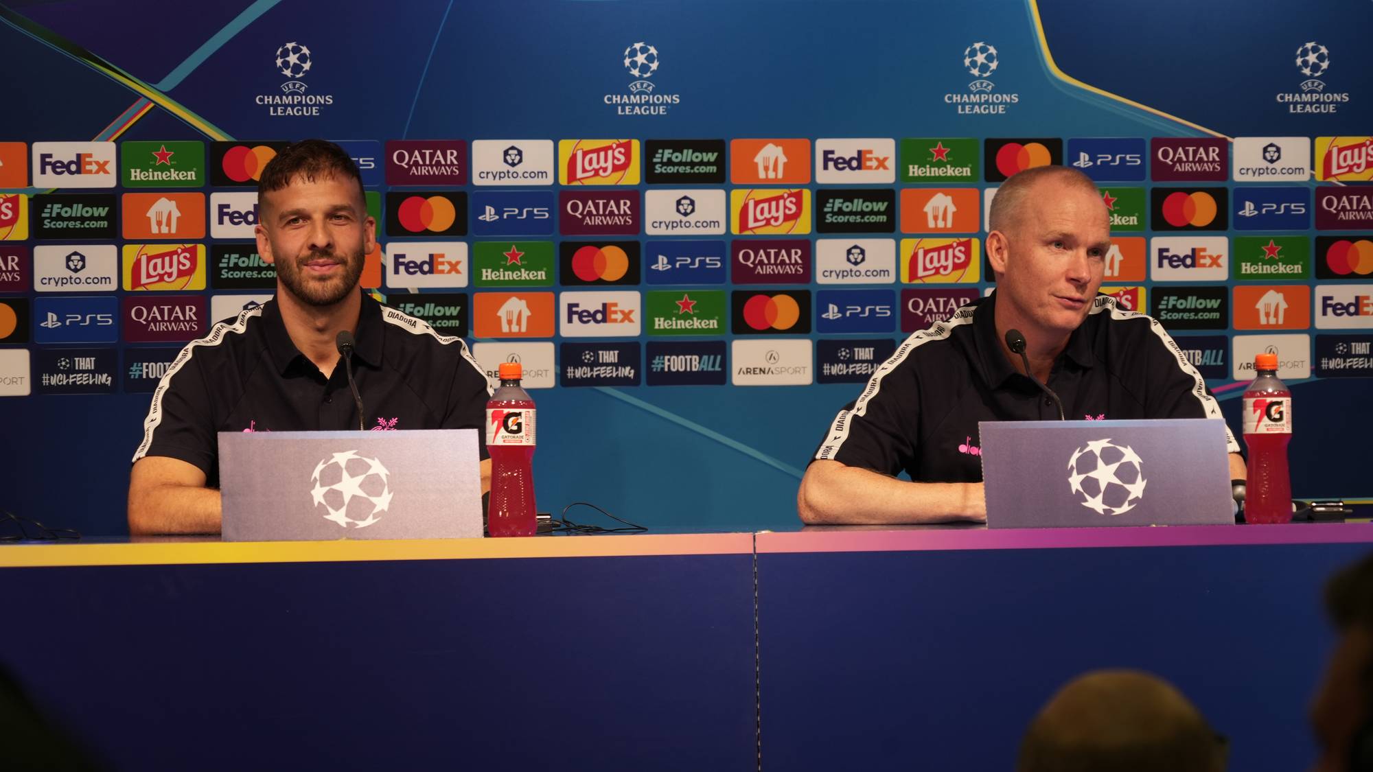 To menn sitter ved et bord foran en pressevegg med Champions League-logoer. (Bildebeskrivelsen er laget av en KI-tjeneste)