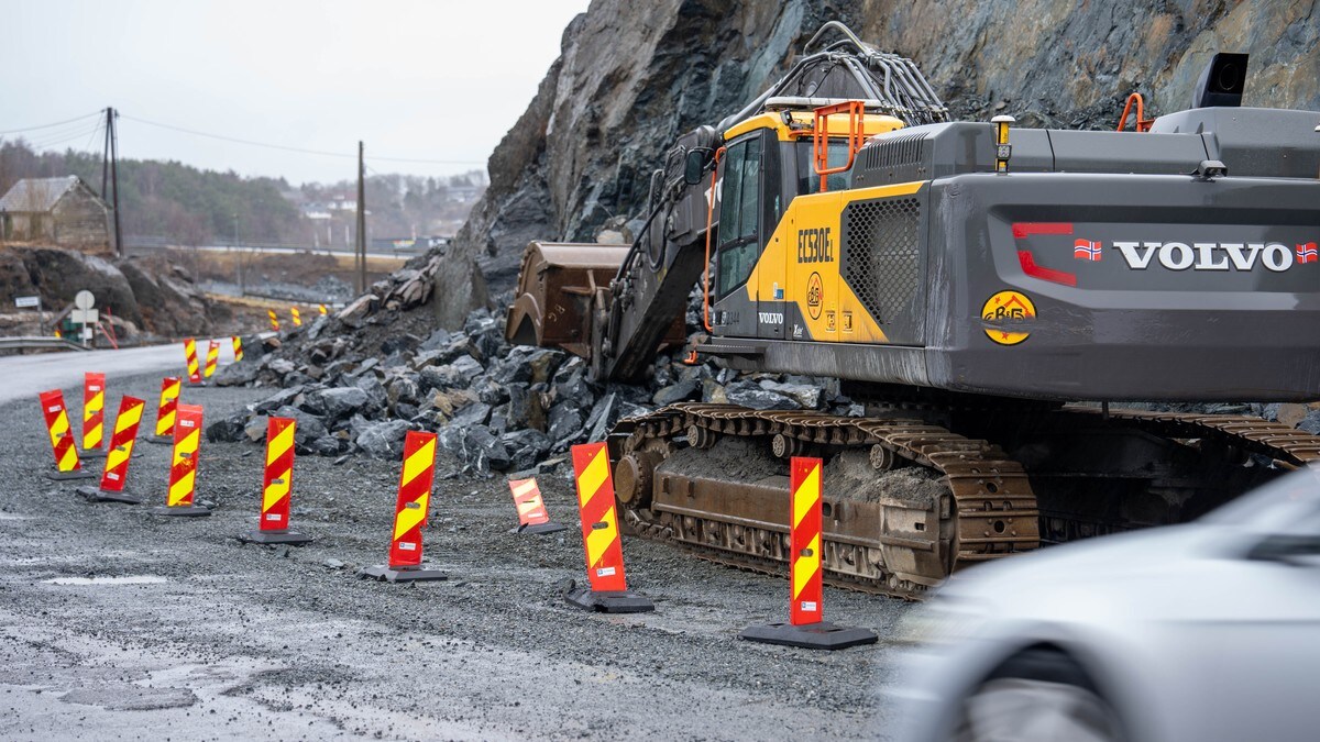 En gravemaskin står i veikant ved en steinbrudd i et byggeområde. Røde og gule skilte markerer avstengte områder rundt maskinen. En grå bil passerer samtidig i veibanen. Bakgrunnen viser en ufullendt vei og noen bygninger i det fjerne. (Bildebeskrivelsen er laget av en KI-tjeneste) FOTO: Olav Røli/NRK