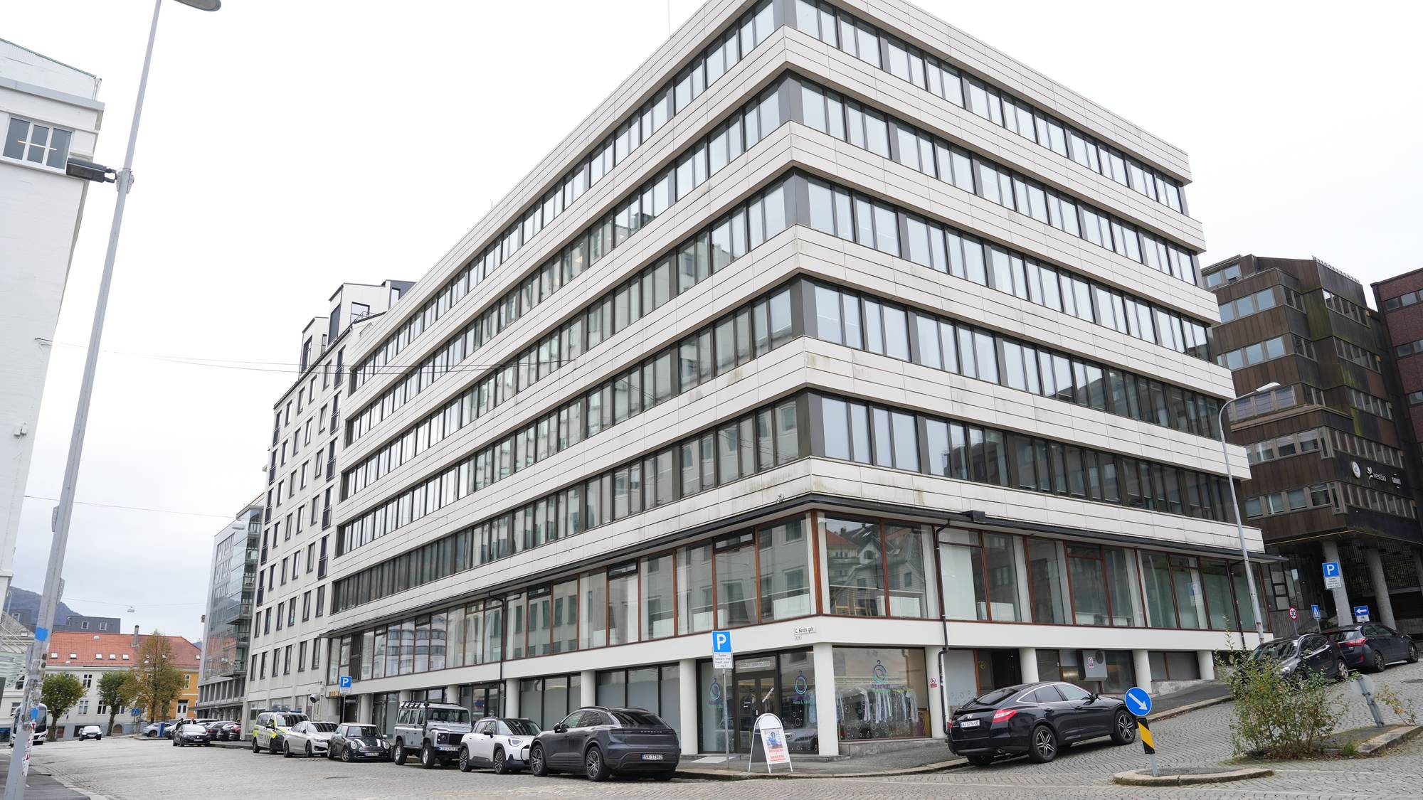 Et moderne kontorbygg i flere etasjer er avbildet på en gate med brosteinsbelagte arbeidsflaten. Flere biler står parkert langs bygningens fasade. Omgivelsene inkluderer lavere bygninger og trær i bakgrunnen, under en grå himmel. Gatebelysning og trafikkskilt er også synlige i bildet.
