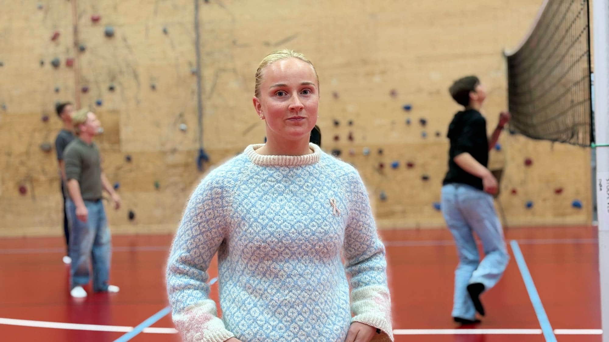 En kvinne med lyst hår i strikk og blå og hvit strikkegenser står i en idrettshall. I bakgrunnen spiller noen gutter volleyball.
