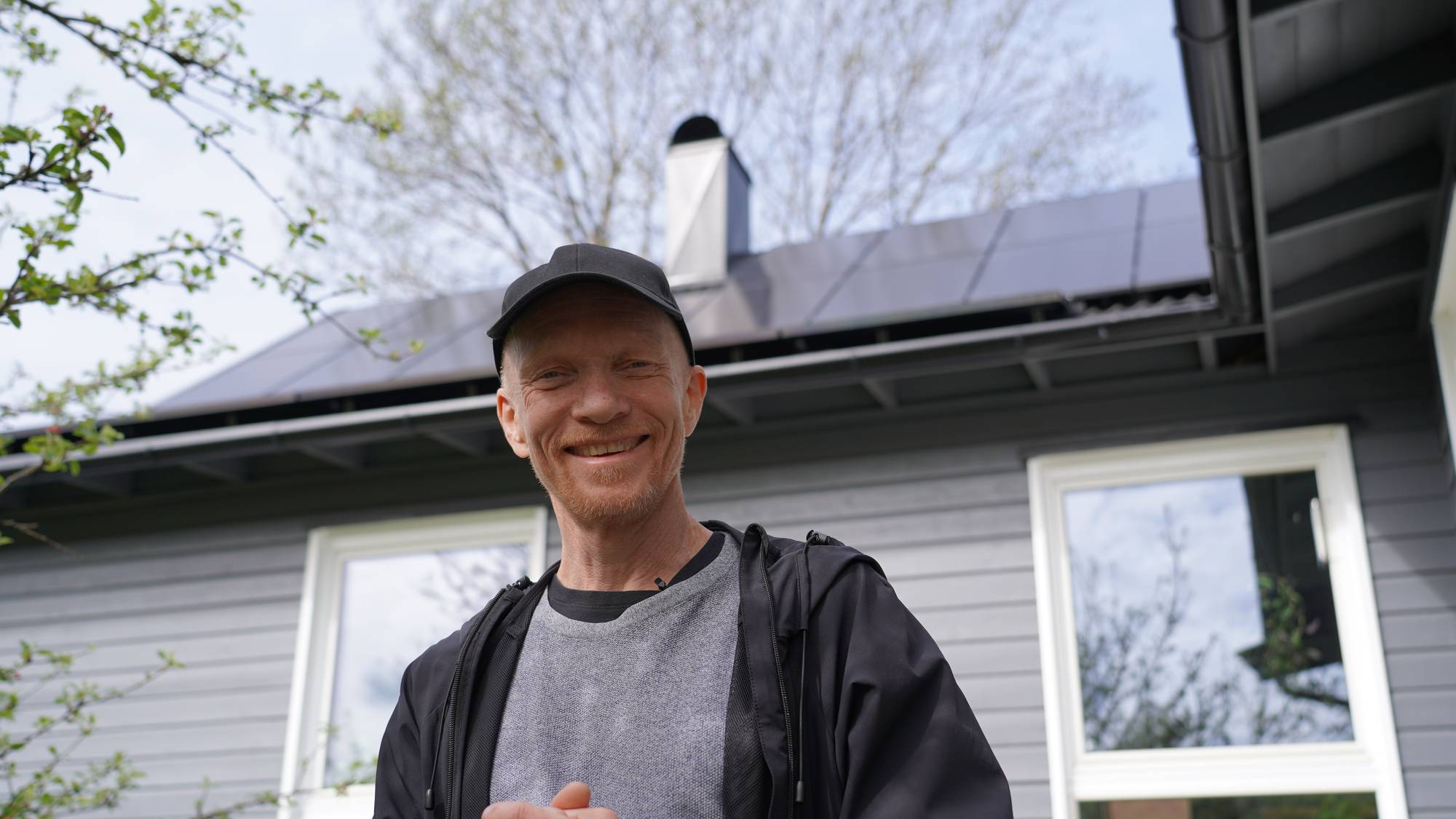Mann med caps står foran et grått hus med solceller på taket. Grønne grener fra et tre til venstre.