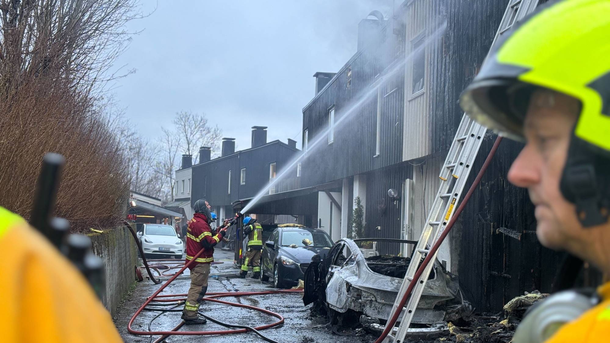 Brannvesen jobber med å slukke en brann i et rekkehus på Voss. En av dem spyler et sort rekkehus med slange. Det stiger røyk fra huset. En bil er utbrent.