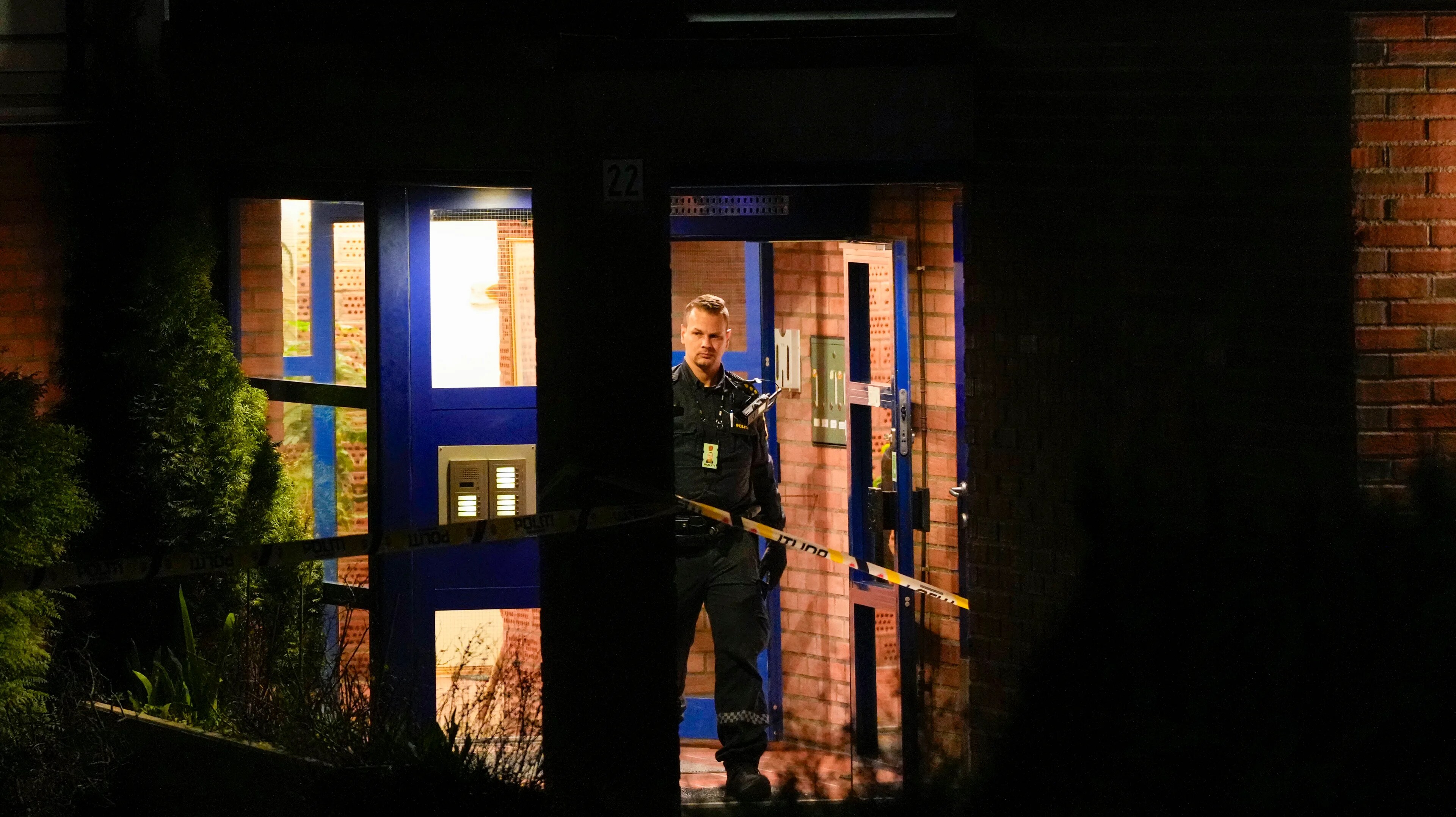 Politibetjent står i inngangen til en boligblokk sperret av med politisperrebånd. (Bildebeskrivelsen er laget av en KI-tjeneste)