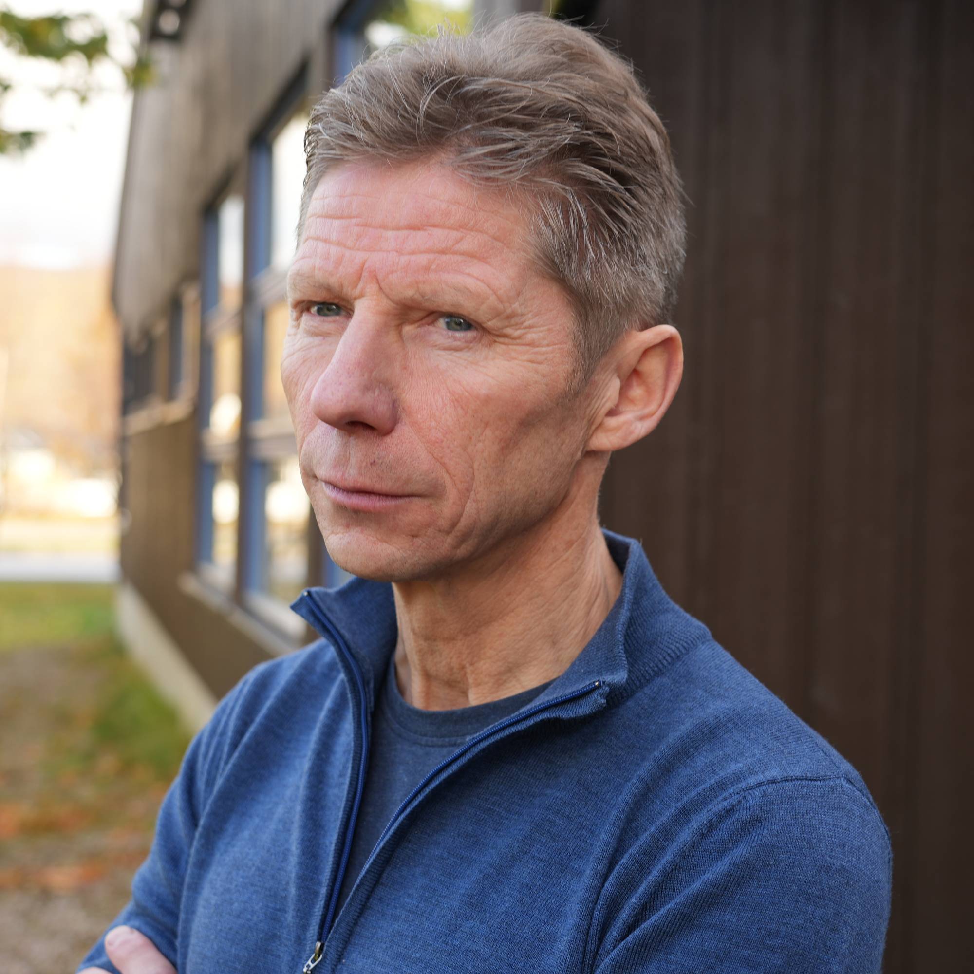 Portrett av en eldre mann med grått hår i en blå genser, stående utendørs ved en trevegg. (Bildebeskrivelsen er laget av en KI-tjeneste)