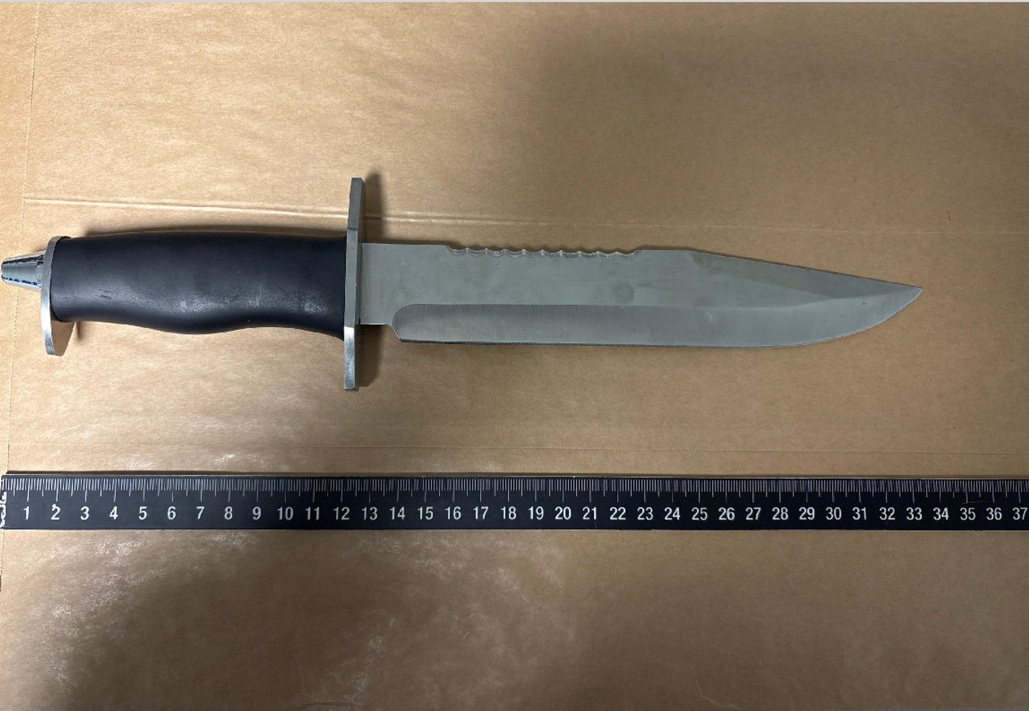 Et stort kniv med svart håndtak og et langt, bredt blad ligger på en brun overflate. Kniven har en taggete del på oversiden av bladet. En målestokk ligger under kniven, som viser centimeter opp til 37. (Bildebeskrivelsen er laget av en KI-tjeneste)