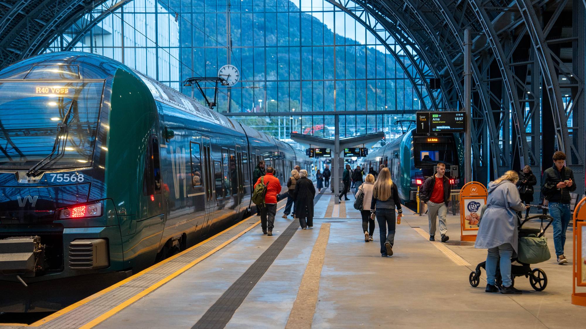 Reisende på perrongen ved Bergen stasjon med tog på begge sider under det buede taket. (Bildebeskrivelsen er laget av en KI-tjeneste)