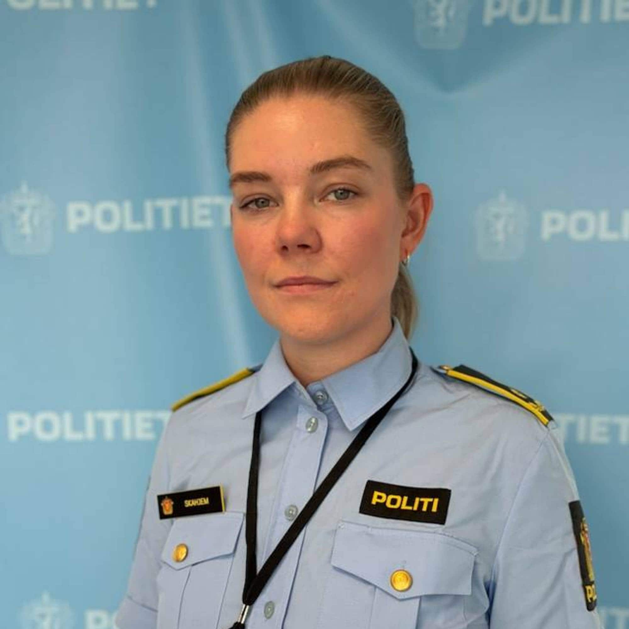 Portrett av en kvinne i politiuniform foran en blå bakgrunn med politilogoer. (Bildebeskrivelsen er laget av en KI-tjeneste)