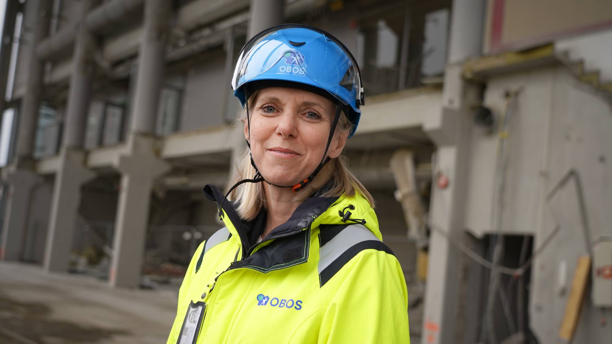 Kvinne med gul vernejakke og blå hjelm står foran et bygg under oppføring. (Bildebeskrivelsen er laget av en KI-tjeneste)
