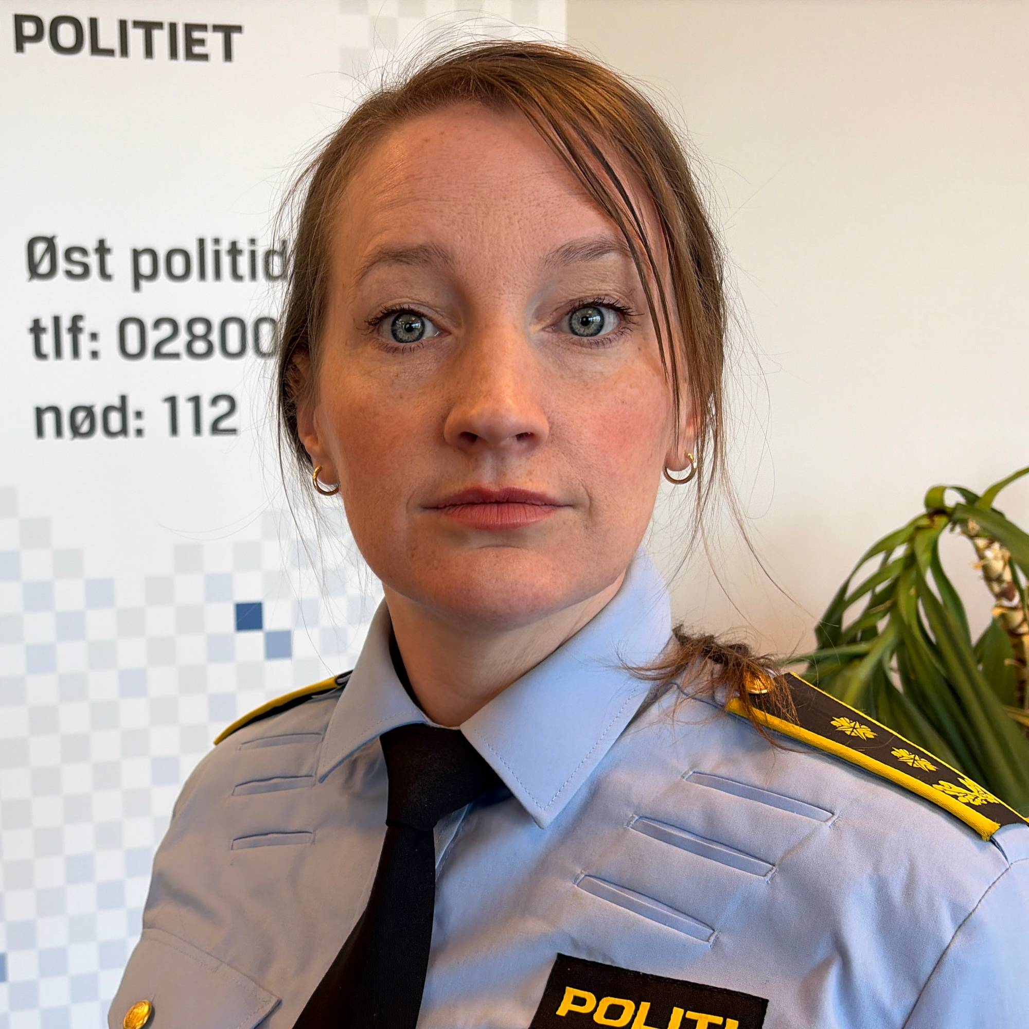 En kvinne i politiuniform står foran en reklameplakat for Øst politidistrikt. Hun har mørkt hår og ser mot kamera. I bakgrunnen er en blå vegg og en potteplante. Plakaten har tekst med telefonnummer og nødnummer. (Bildebeskrivelsen er laget av en KI-tjeneste)