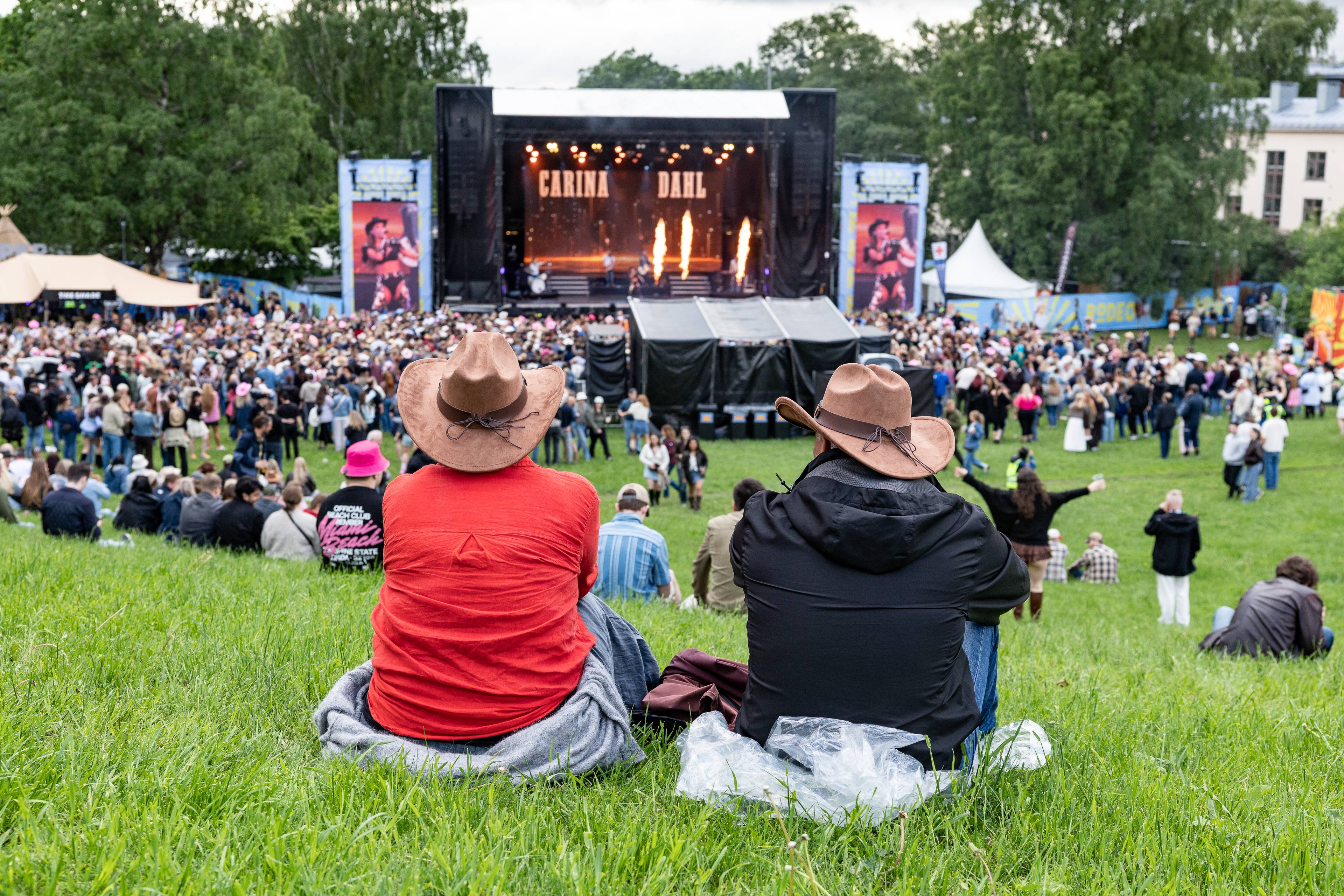 To personer med cowboyhatt sitter på en gresslette og ser mot en scene. Publikum samler seg foran scenen der en artist, Carina Dahl, opptrer. Det er et stort utendørs arrangement med flere telt og fasiliteter. (Bildebeskrivelsen er laget av en KI-tjeneste)