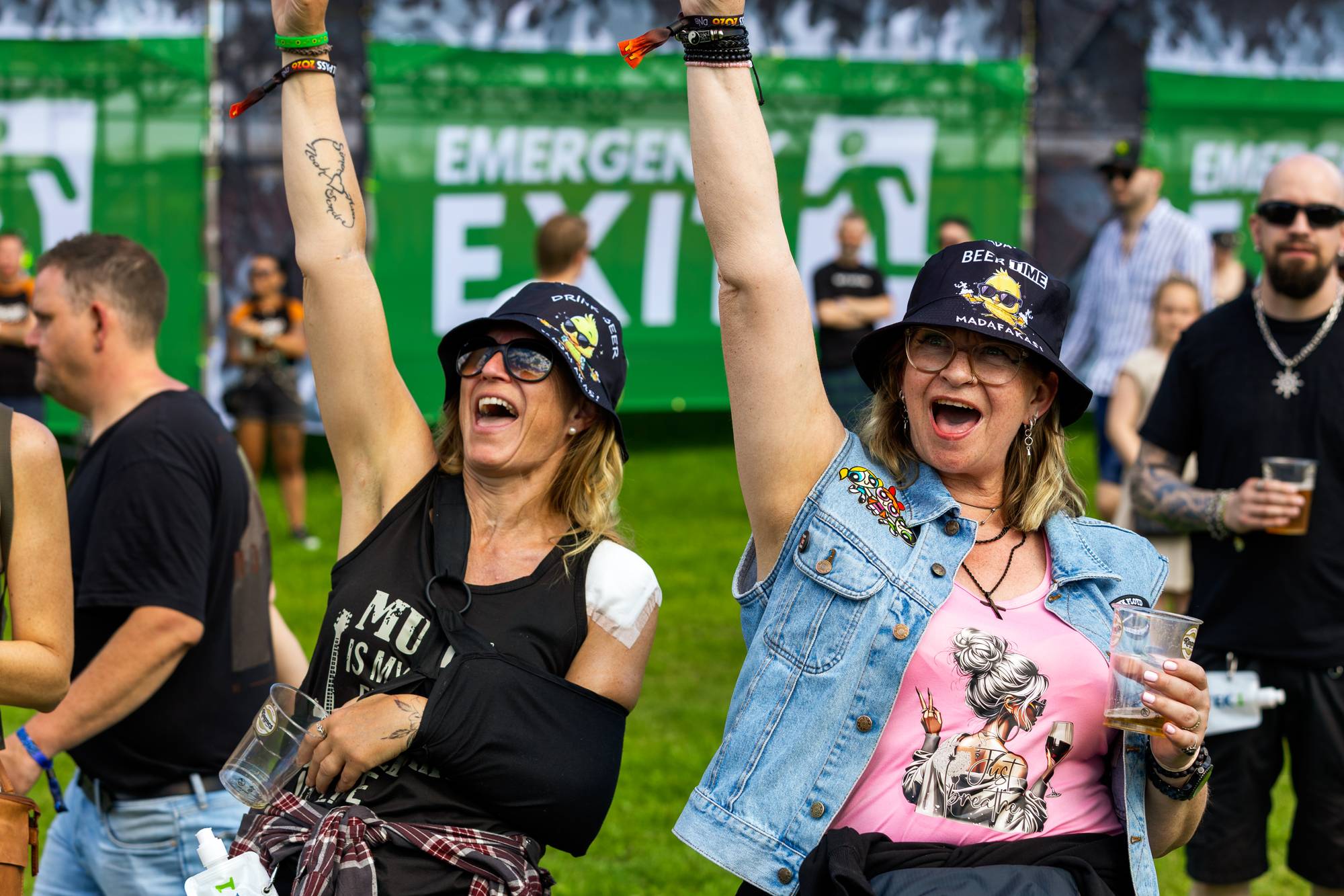 To kvinner med armene hevet heier på en festival. Begge har på seg hatt og ser ut til å synge eller rope. Bak dem er det folkemengde og en grønn plakat med "EMERGENCY EXIT". (Bildebeskrivelsen er laget av en KI-tjeneste)