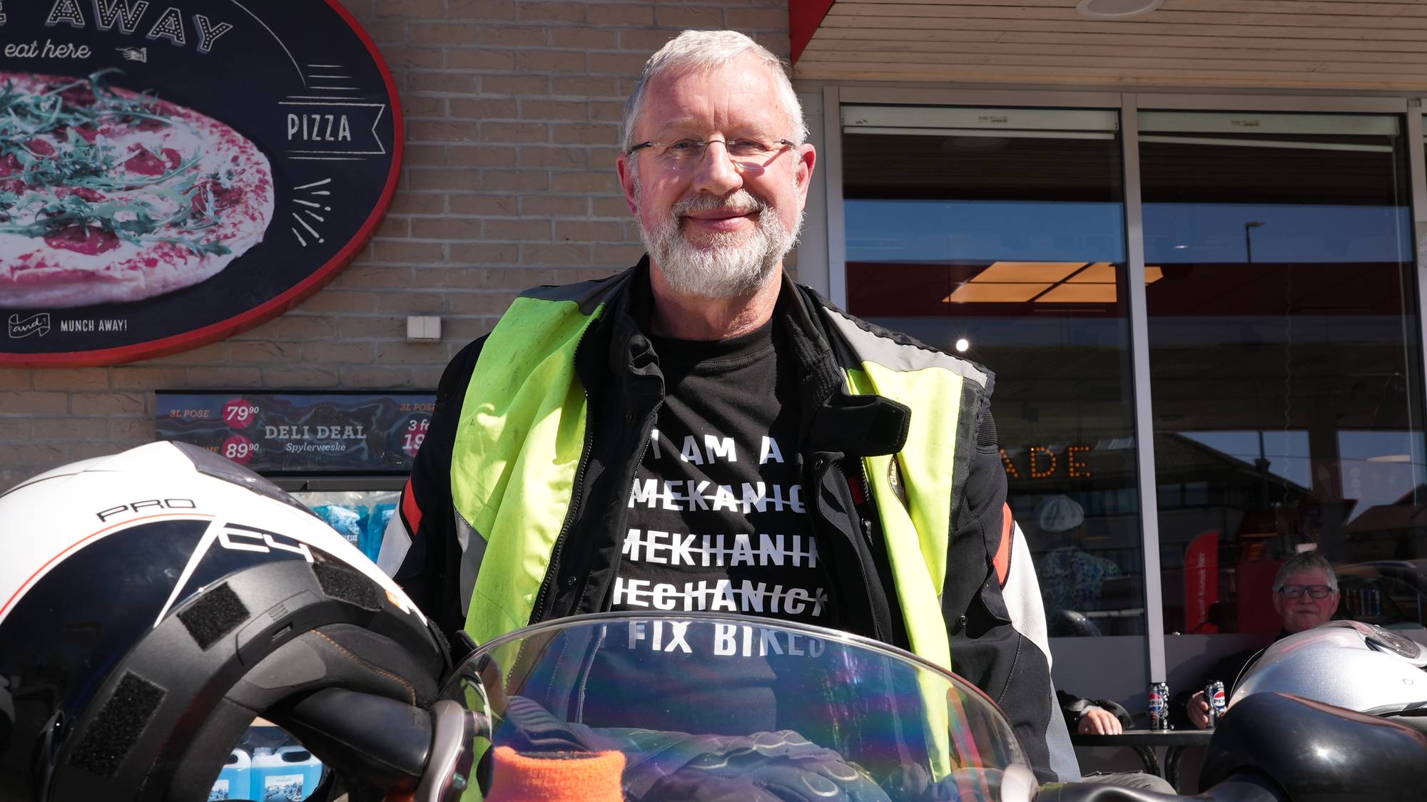En mann med skjegg og briller står foran en motorsykkel. Han har på seg en gul vest over en t-skjorte med tekst. Bak ham er en murvegg med et rundt skilt med pizzabilde og en butikkfasade med vinduer. En hjelm hviler på motorsykkelen foran mannen. En annen person sitter ved et bord inne i butikken. (Bildebeskrivelsen er laget av en KI-tjeneste)