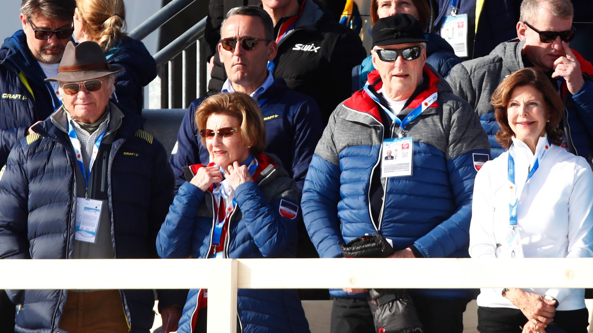 Kong Harald og dronning Sonja sitter på en tribune omgitt av tilskuere under et skirenn. 