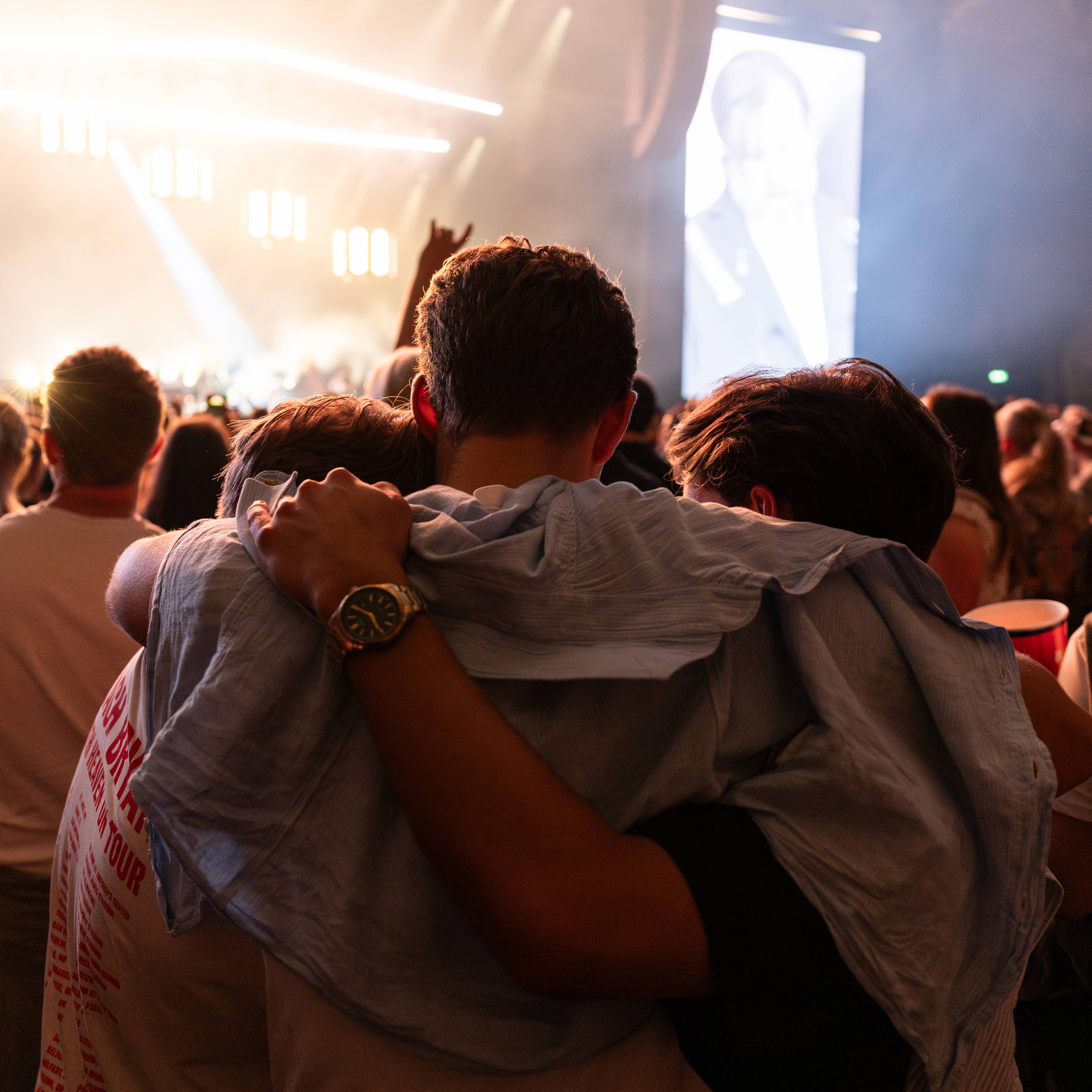 Publikum ser på en scene under en konsert. Tre personer i forgrunnen omfavner hverandre. En stor skjerm viser en person i bakgrunnen. Lys og røyk skaper en dramatisk stemning. (Bildebeskrivelsen er laget av en KI-tjeneste)
