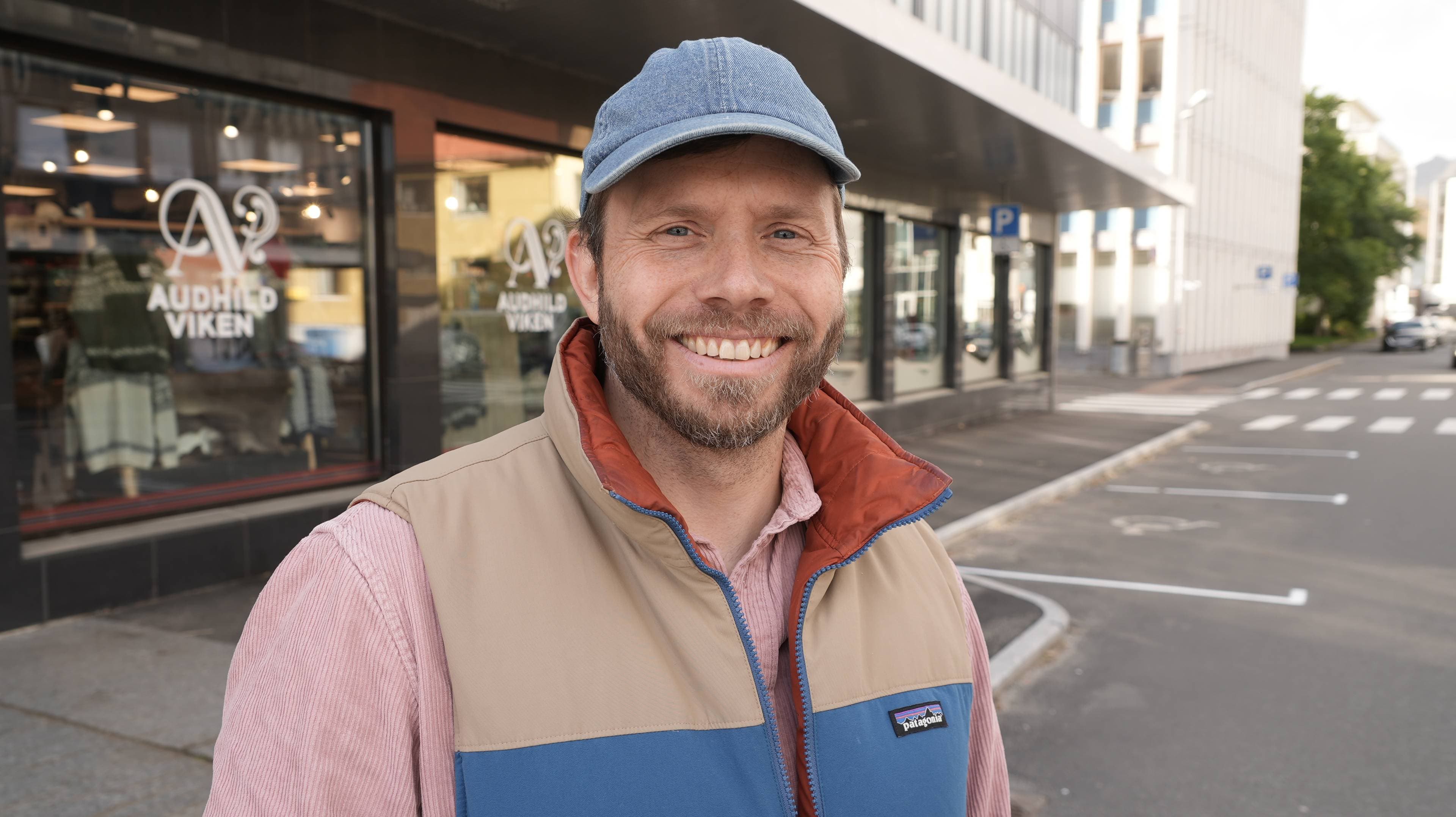 En mann med beige og blå fleacevest over en rosa skjorte og caps står på en parkeringsplass foran et butikkvindu og smiler til kamera. Han har brunt hår og skjegg.
