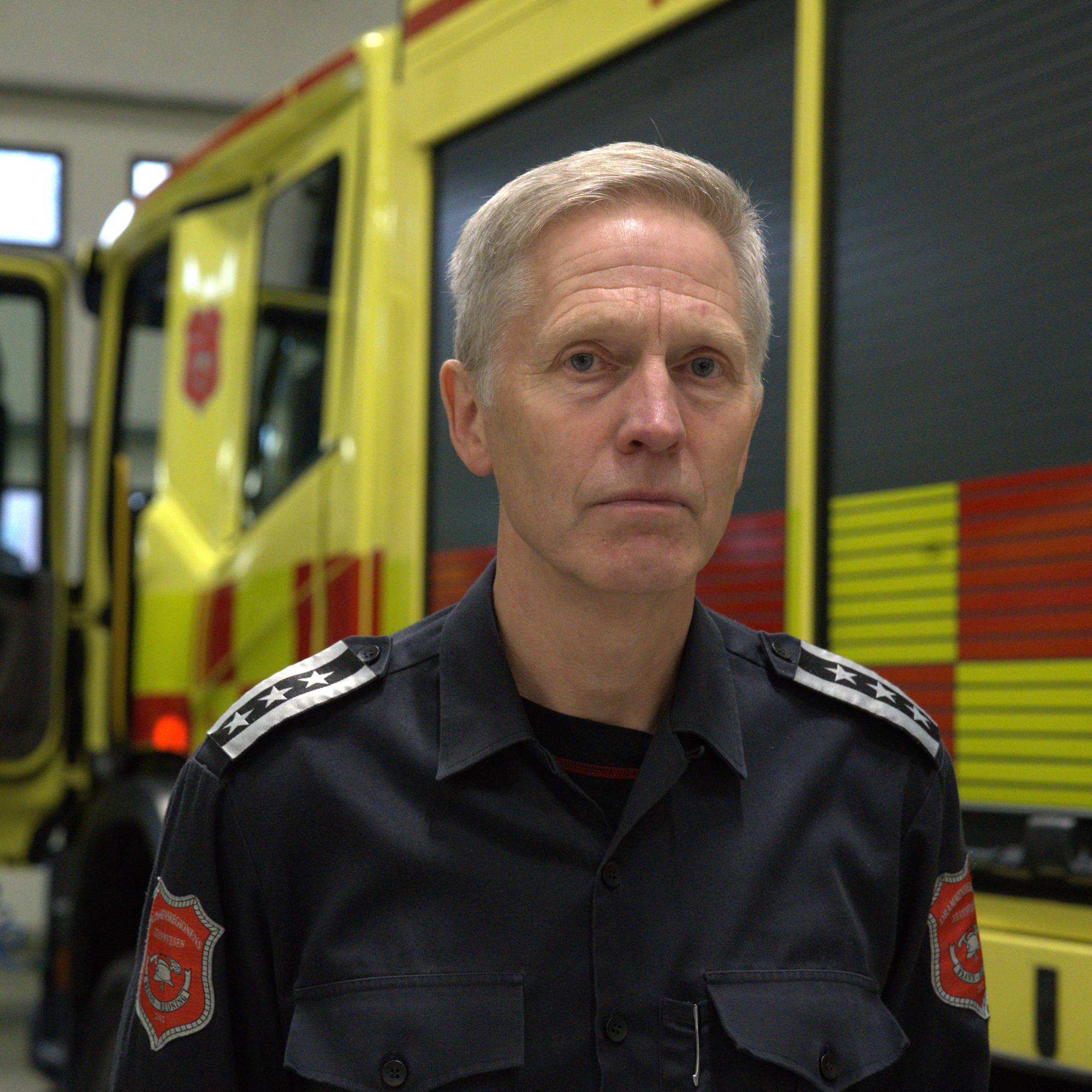 Brannsjefen i Drammen, Torgeir Andersen ser seriøst mot kamera. Han har kort hvitt hår. Han står foran en brannbil. 
