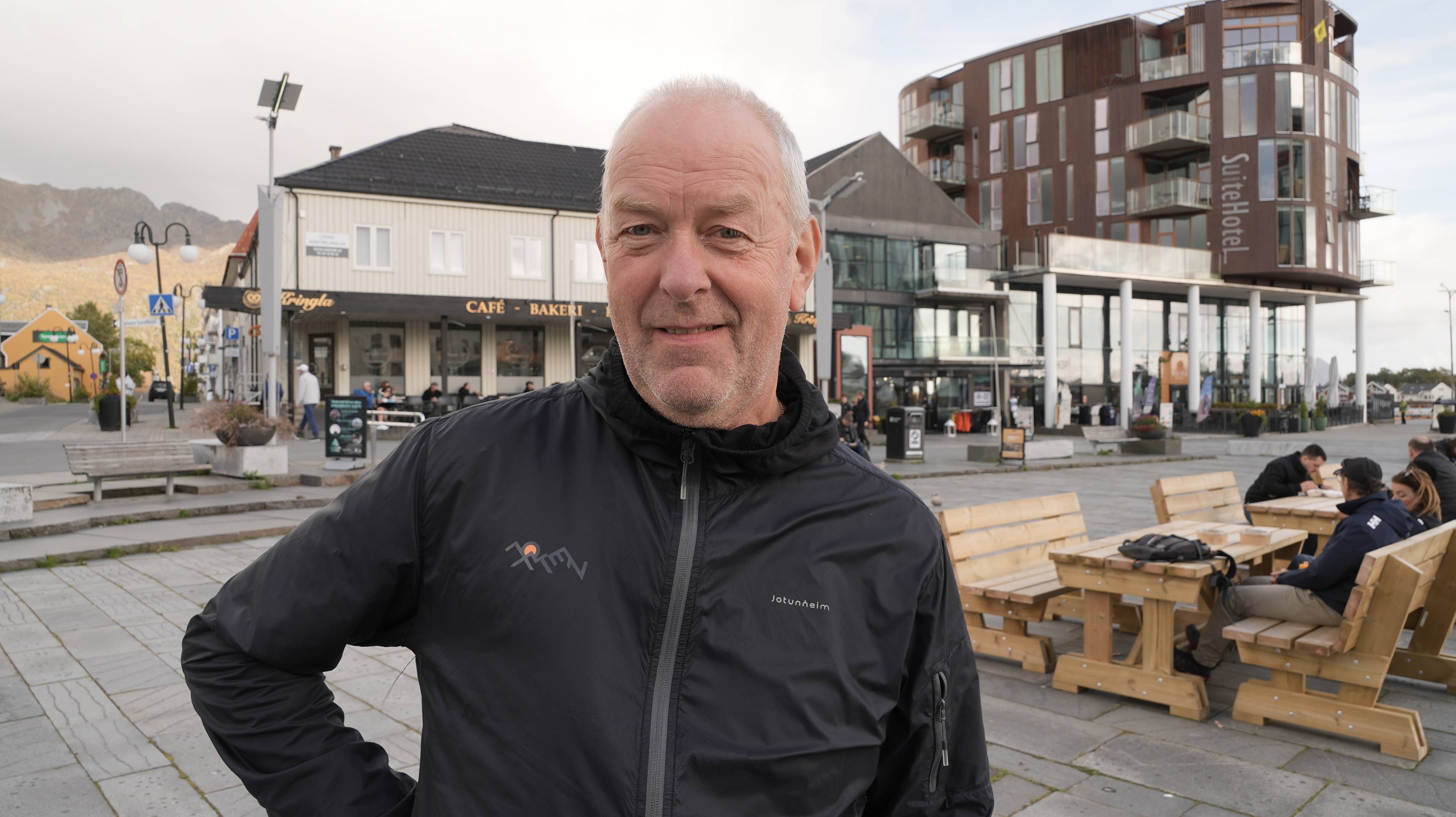 En mann med kort grått hår og skjegg og svart jakke ser mot kamera. I bakgrunn sitter folk på trebenker og slapper av.