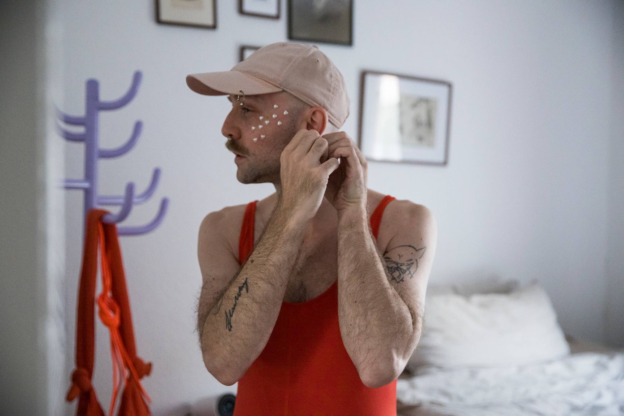 En mann med beige caps og rød singlet. Han har paljetter i ansiktet og tatovering på armene, og setter i en øredobb. 
