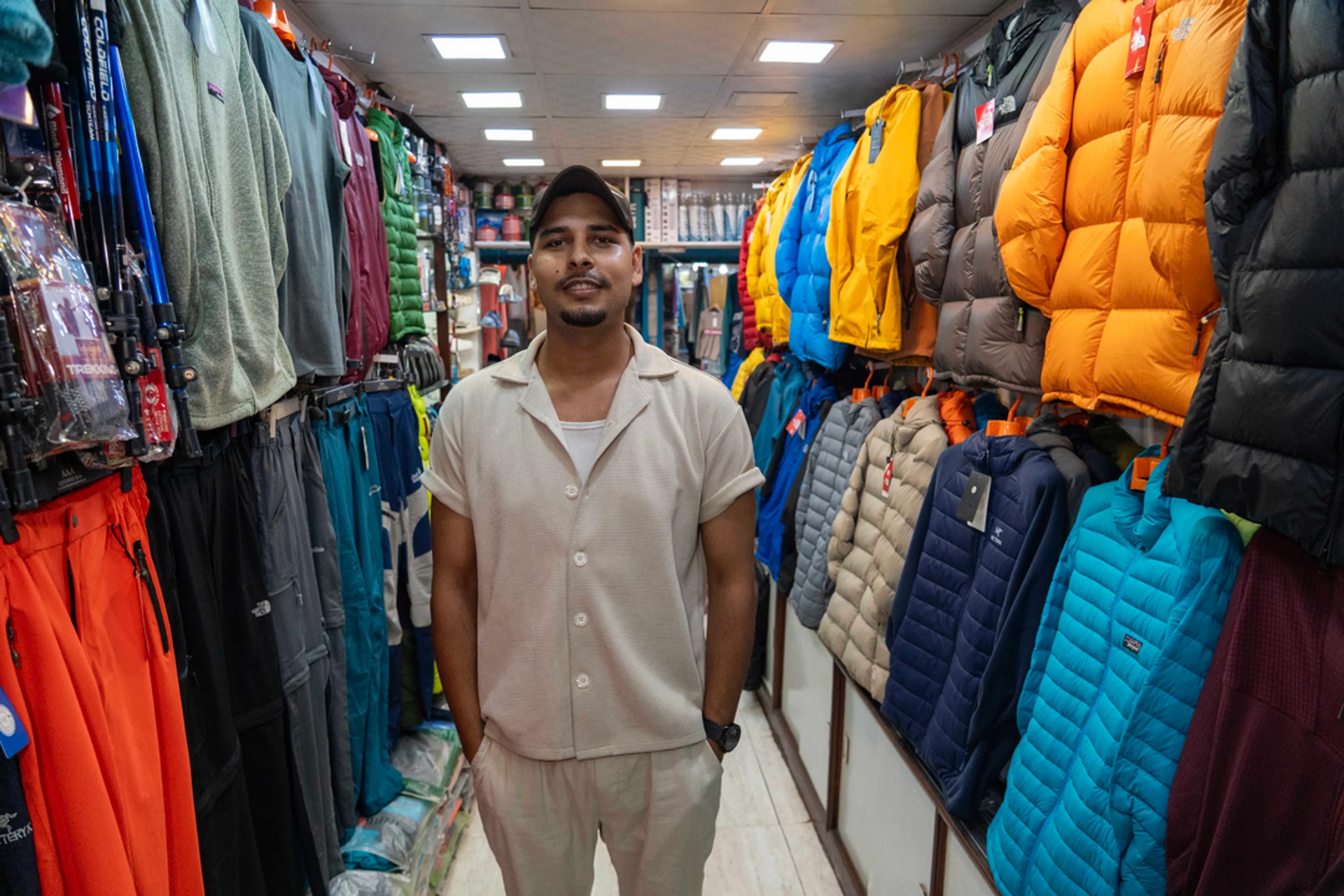 Sel utstyr i Katmandu til turistar som skal til fjells – Ein mørk mann i lyst tøy står mellom to rader av fargerike dunjakker og anna sportsutstyr