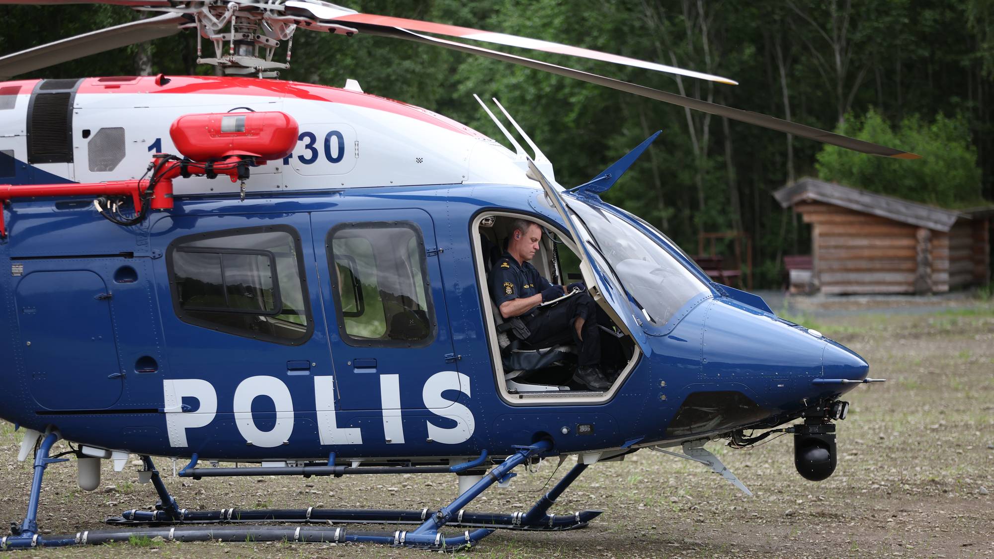 Politibetjent i et svensk politihelikopter på bakken ved en skog. (Bildebeskrivelsen er laget av en KI-tjeneste)