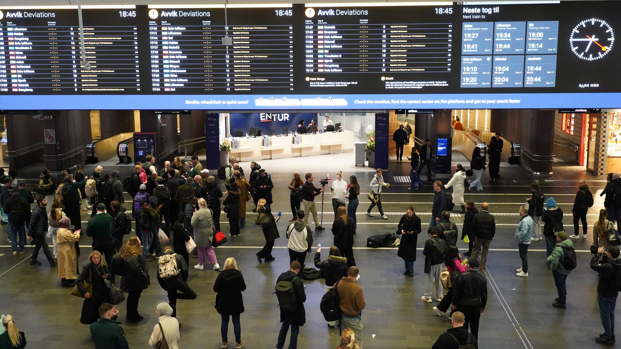 Reisende samles i en togstasjonshall under en storskjerm som viser avvik i togtrafikken. (Bildebeskrivelsen er laget av en KI-tjeneste)